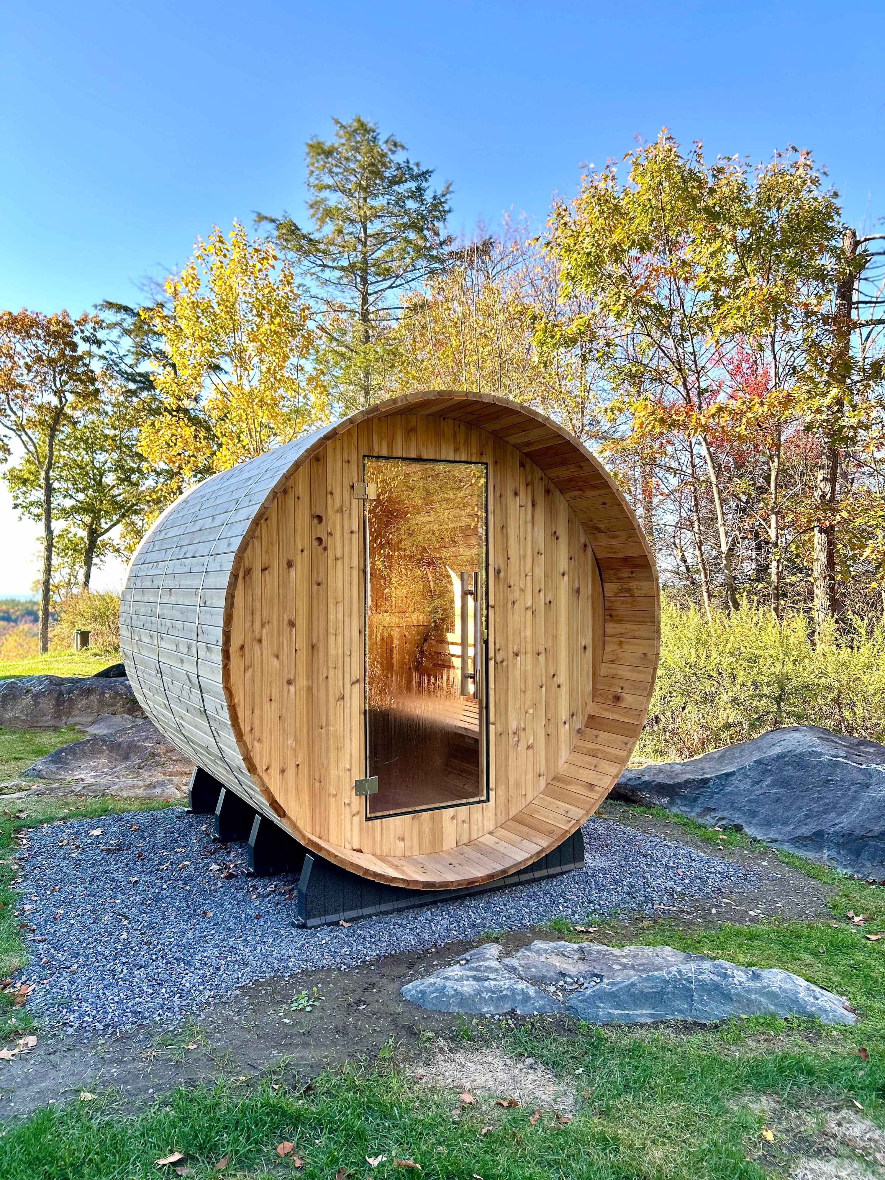 Outdoor barrel-shaped sauna made of light wood with a glass door, set on a gravel base amidst rocks and green grass, surrounded by trees.