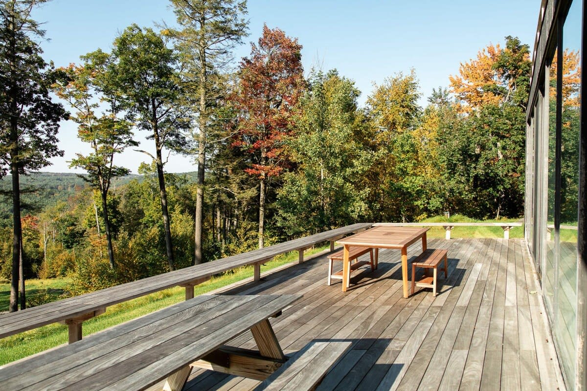 Expansive wooden deck furnished with a large picnic table and long benches, offering panoramic views of a dense, colorful forest under a clear blue sky, ideal for outdoor dining and relaxation.