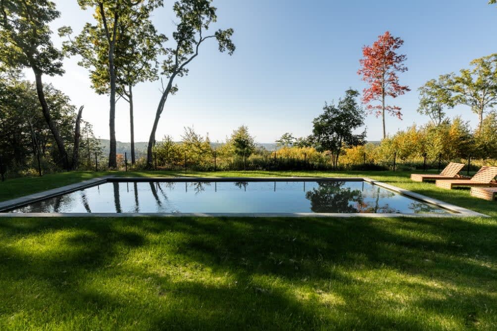 A wide shot of the rectangular swimming pool surrounded by a manicured green lawn, with wooden lounge chairs visible. Tall trees and distant hills with autumn colors frame the serene outdoor scene.