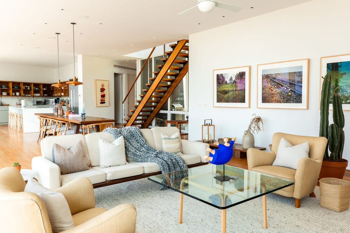 Open-concept living space with a white sofa, beige armchairs, a glass coffee table, and a prominent wooden staircase leading to the upper floor, with the kitchen and dining area visible in the background, promoting a fluid layout.