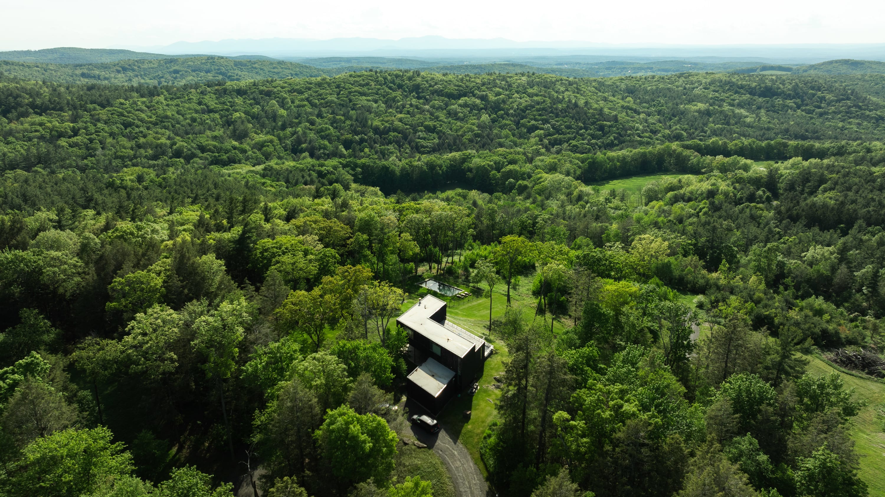 An aerial view showcases the modern, dark-roofed house nestled within a vast, verdant forest, with a winding driveway leading to the property. Distant mountains are visible under a clear sky, emphasizing the secluded location.