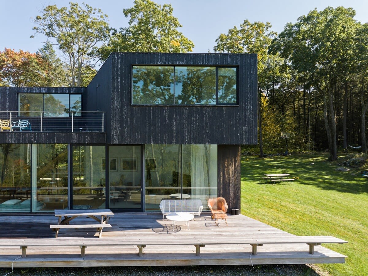 Modern black-clad house with large windows and a spacious wooden deck featuring outdoor seating and a picnic table, set against a lush green lawn and dense forest, offering a blend of contemporary design and natural surroundings.