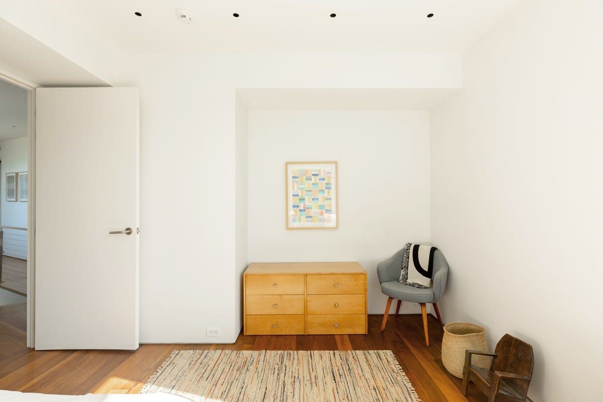 A bright bedroom with white walls, hardwood floors, and a light wood dresser. A grey armchair with a blanket sits beside a framed abstract artwork, with a striped rug on the floor.