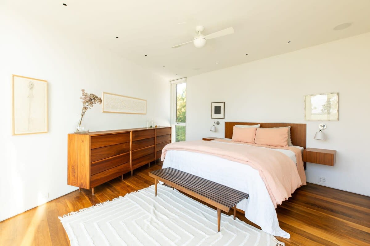 A spacious, sunlit bedroom with rich hardwood floors, a large bed with a wooden headboard and pink bedding, flanked by floating wooden nightstands, and a long wooden dresser against a white wall.