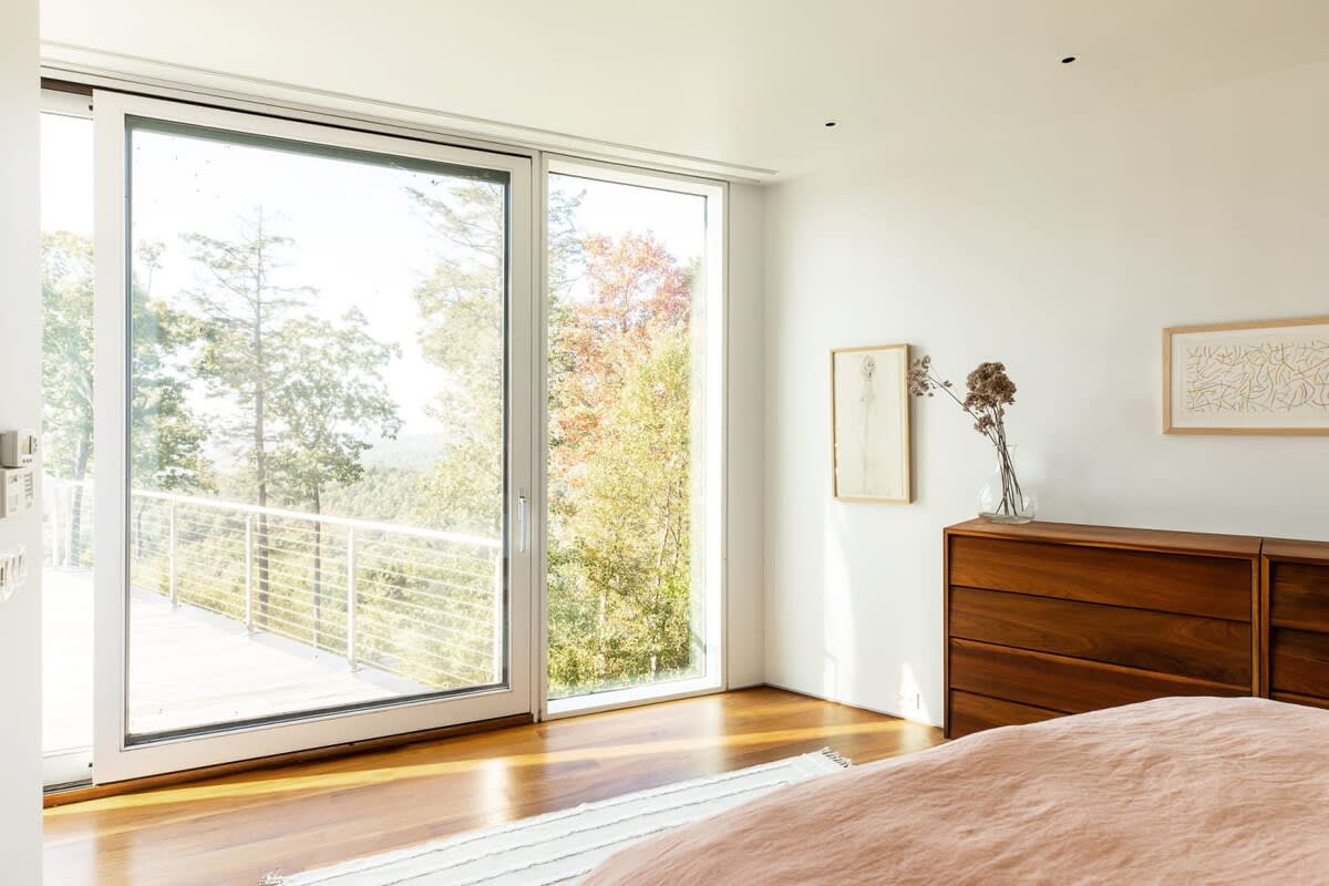 A bright bedroom corner featuring a large sliding glass door and an adjacent tall window, offering expansive views of lush green and autumn-colored trees and a distant landscape.