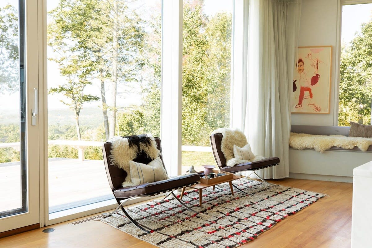 Cozy seating nook with two modern leather chairs adorned with sheepskin throws, a patterned rug, and a small side table, positioned by large windows offering serene outdoor views.