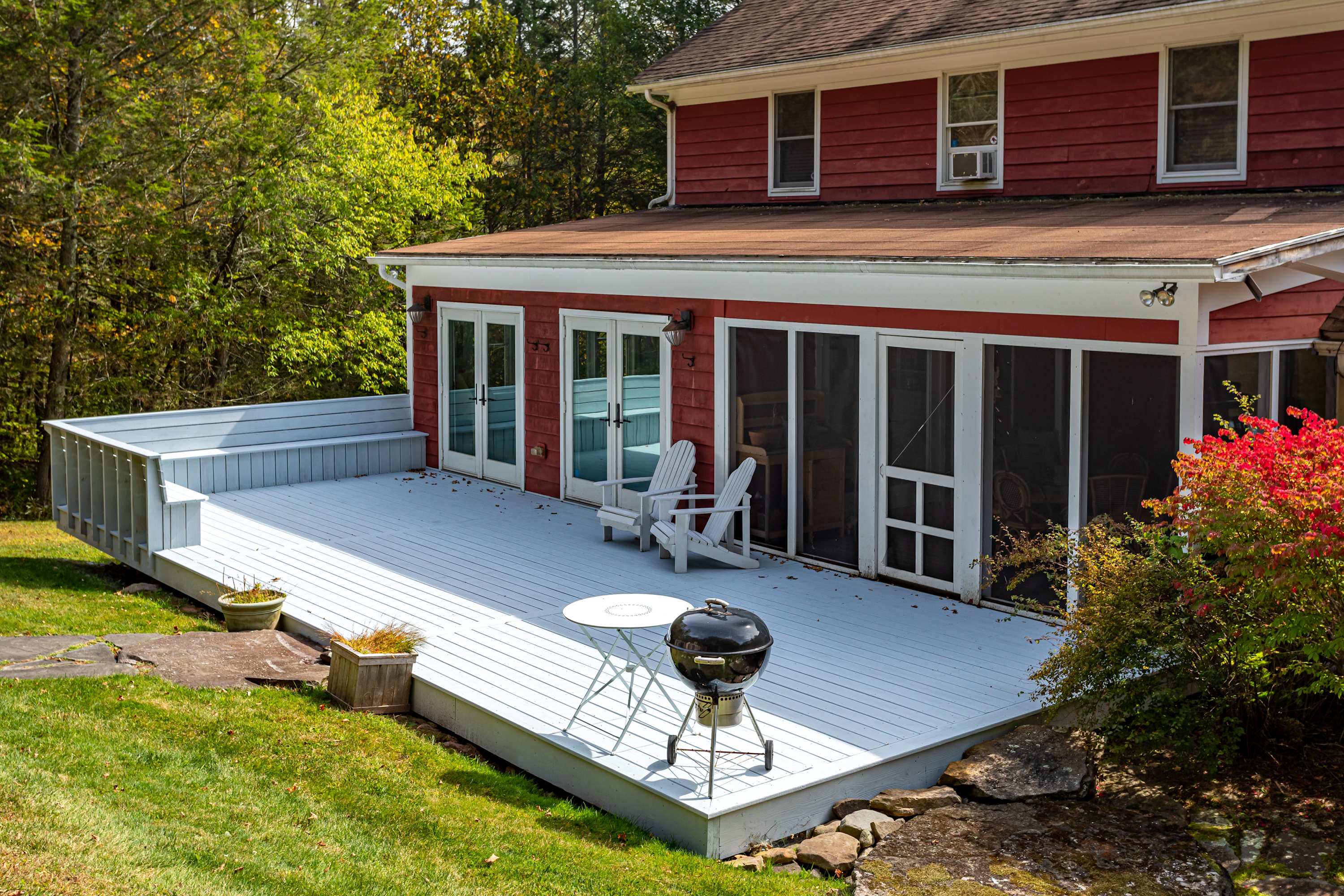 A spacious white wooden deck extends from the red-sided house, featuring two white Adirondack chairs, a small white table, and a black BBQ grill. Lush green grass and trees frame the outdoor space, with multiple glass doors leading into the house.