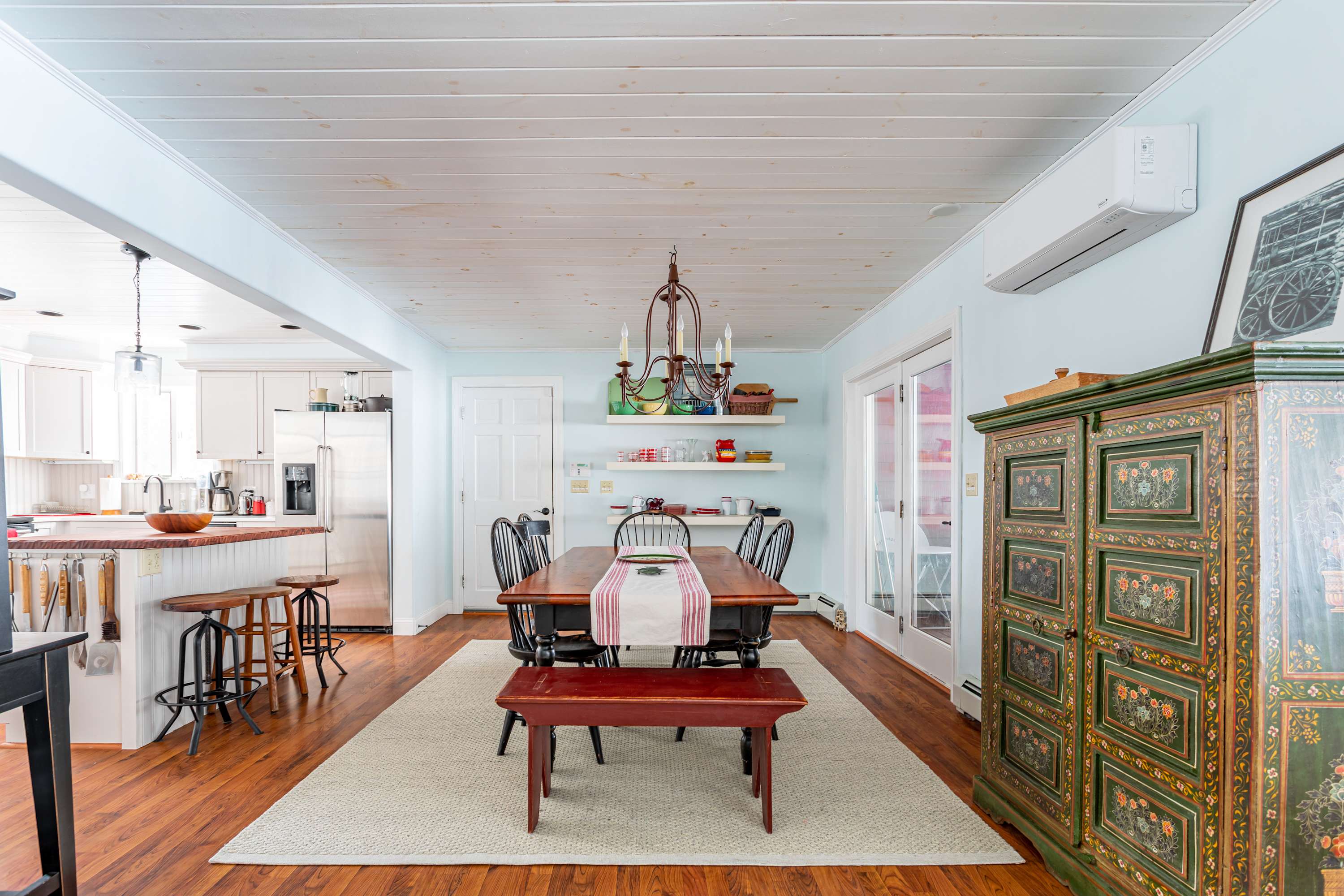 A rustic dining area with a long wooden table, black Windsor chairs, and a red bench, set on a light rug, with a view into the kitchen and living room, featuring a decorative chandelier and light blue walls.