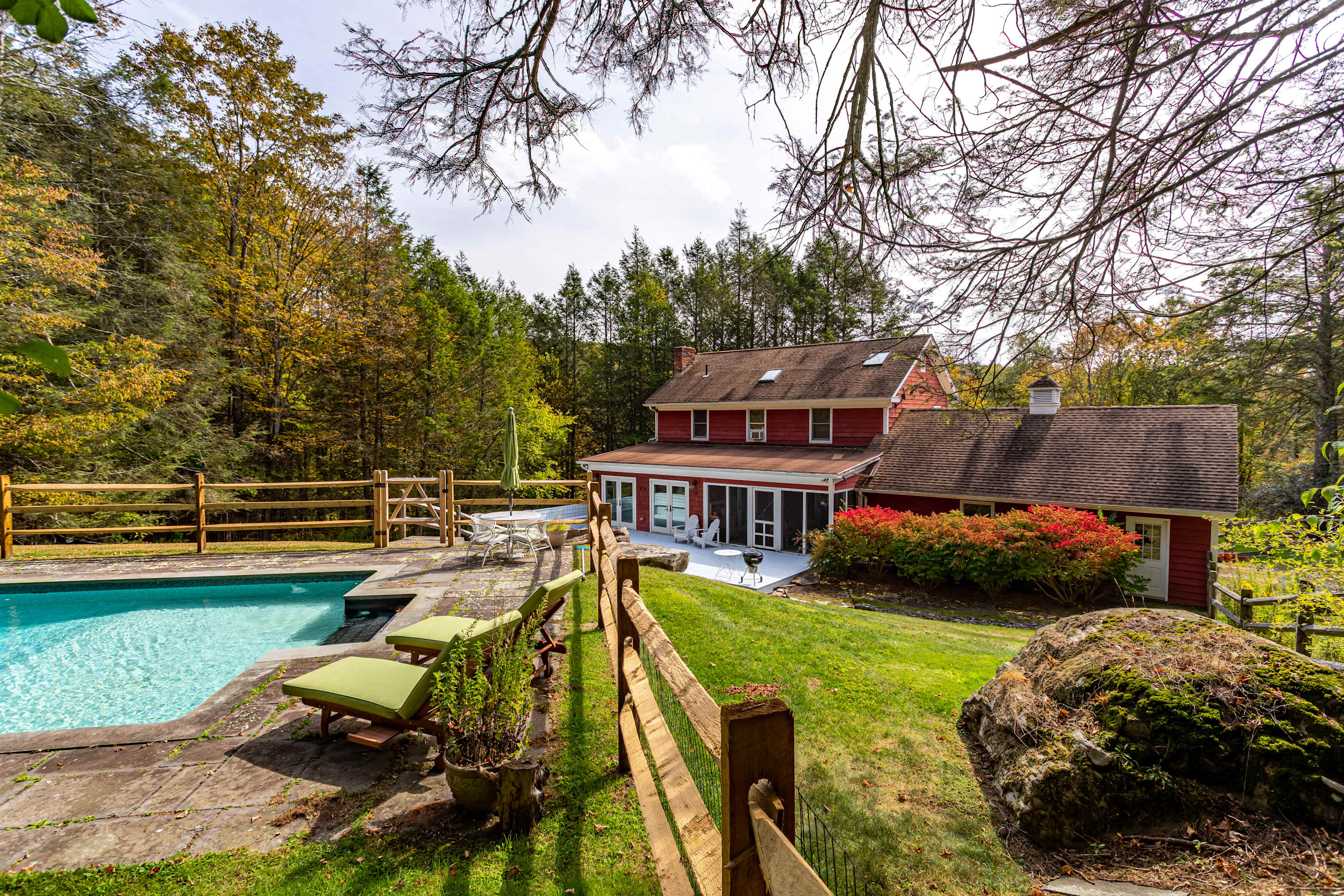 An inviting outdoor scene showcasing a red-sided house with a large swimming pool surrounded by a stone patio and green lawn. A wooden fence encloses the area, with lush trees and foliage in the background under a partly cloudy sky.