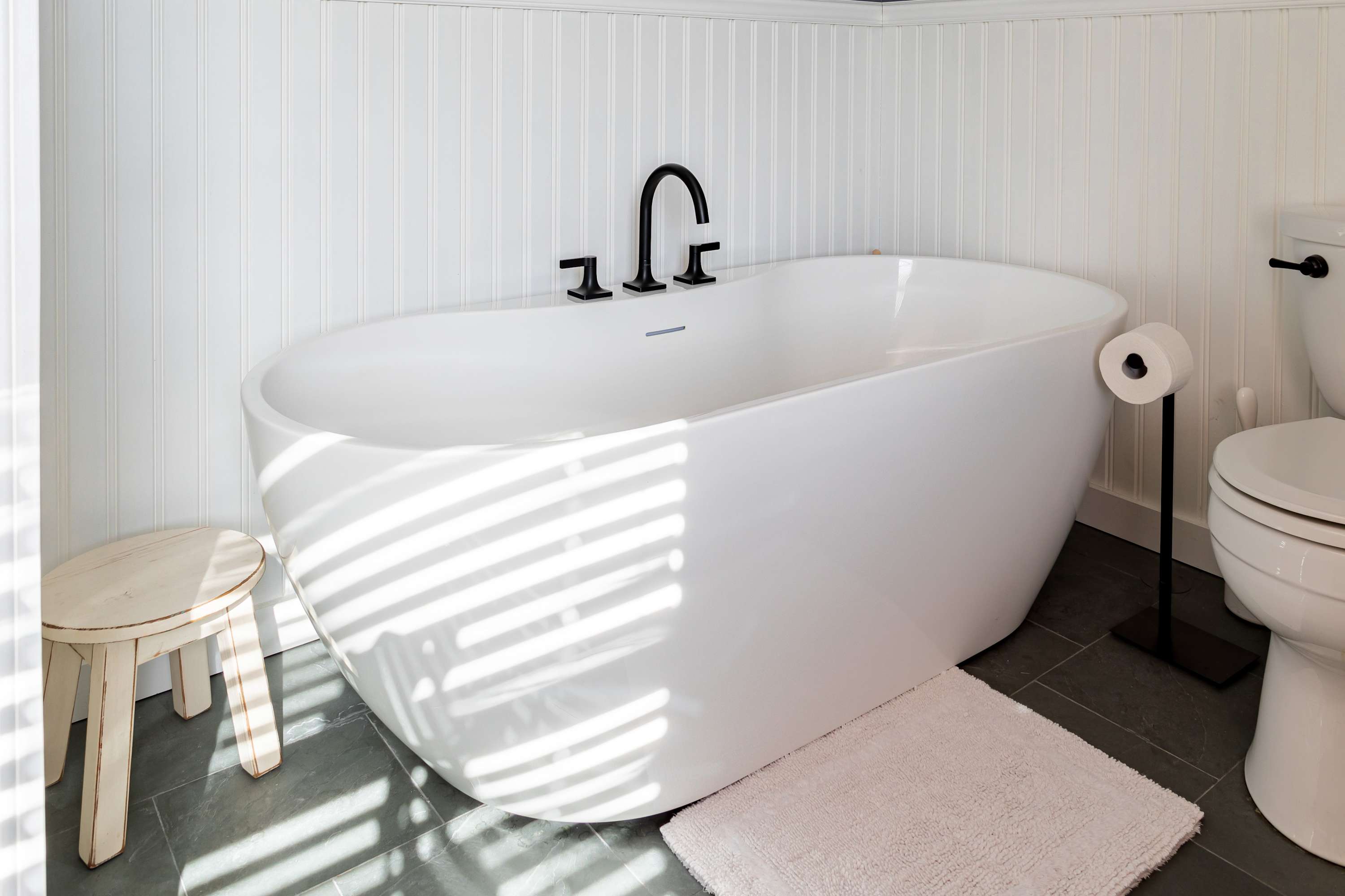 A close-up of a sleek white freestanding soaking tub with modern black fixtures, positioned against white wainscoting, next to a toilet and a small wooden stool, with sunlight streaming through a window.