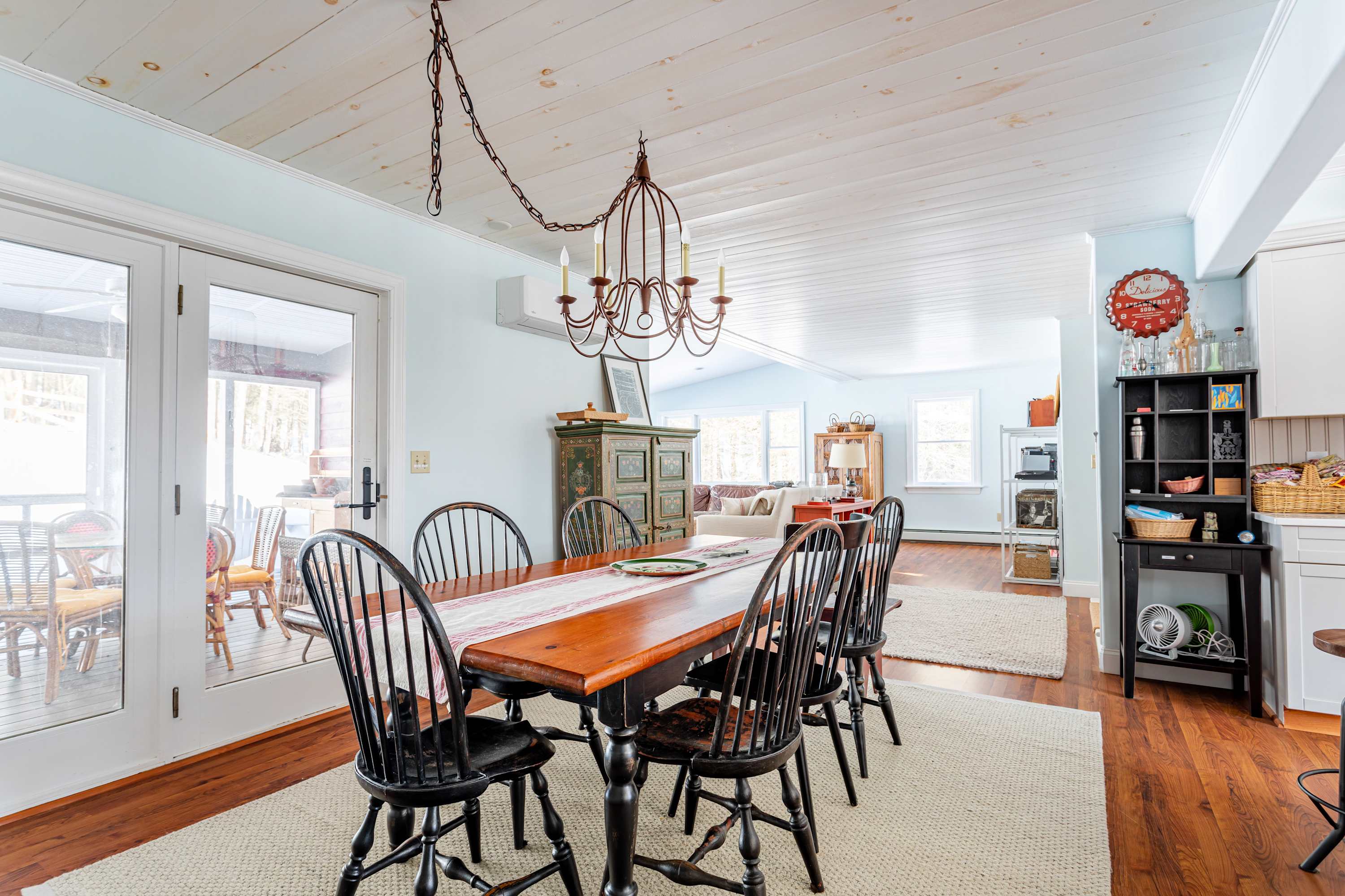 A closer view of the dining area with a long wooden table, black Windsor chairs, and a red bench, showing double glass doors leading to the outside, and a glimpse of the living room beyond.
