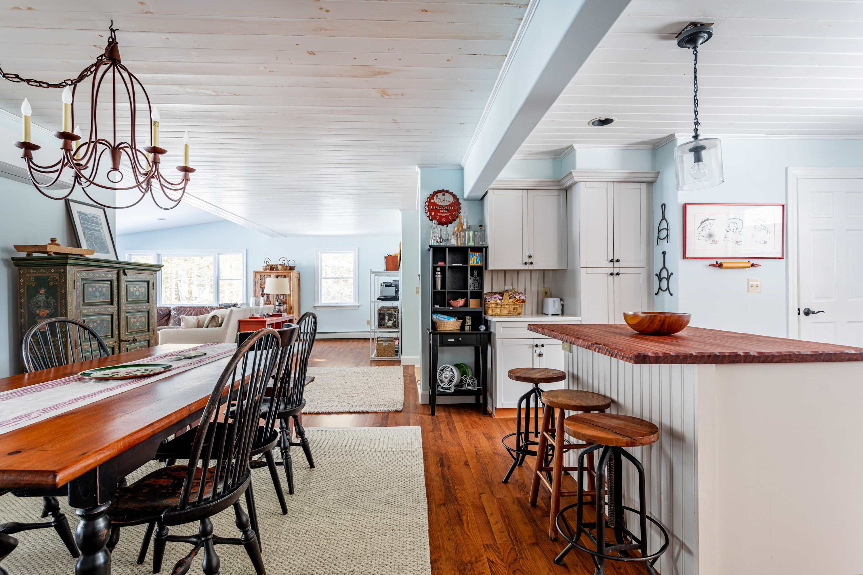 Open concept dining area with a long wooden table and black chairs, leading into a kitchen with white cabinets and a wooden island, all under a white plank ceiling.