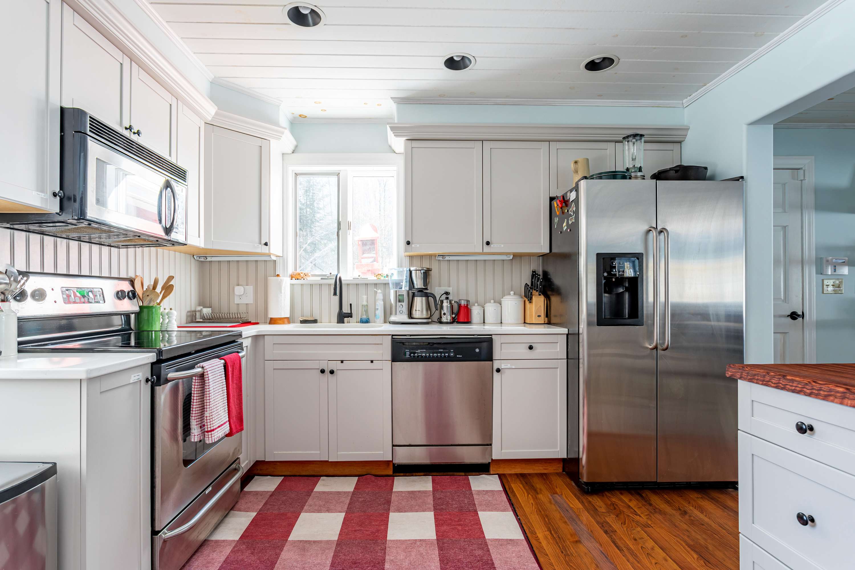 A well-equipped kitchen with light gray shaker cabinets, stainless steel appliances including a refrigerator, oven, and dishwasher, and a window above the sink, featuring a red and white checkered rug.