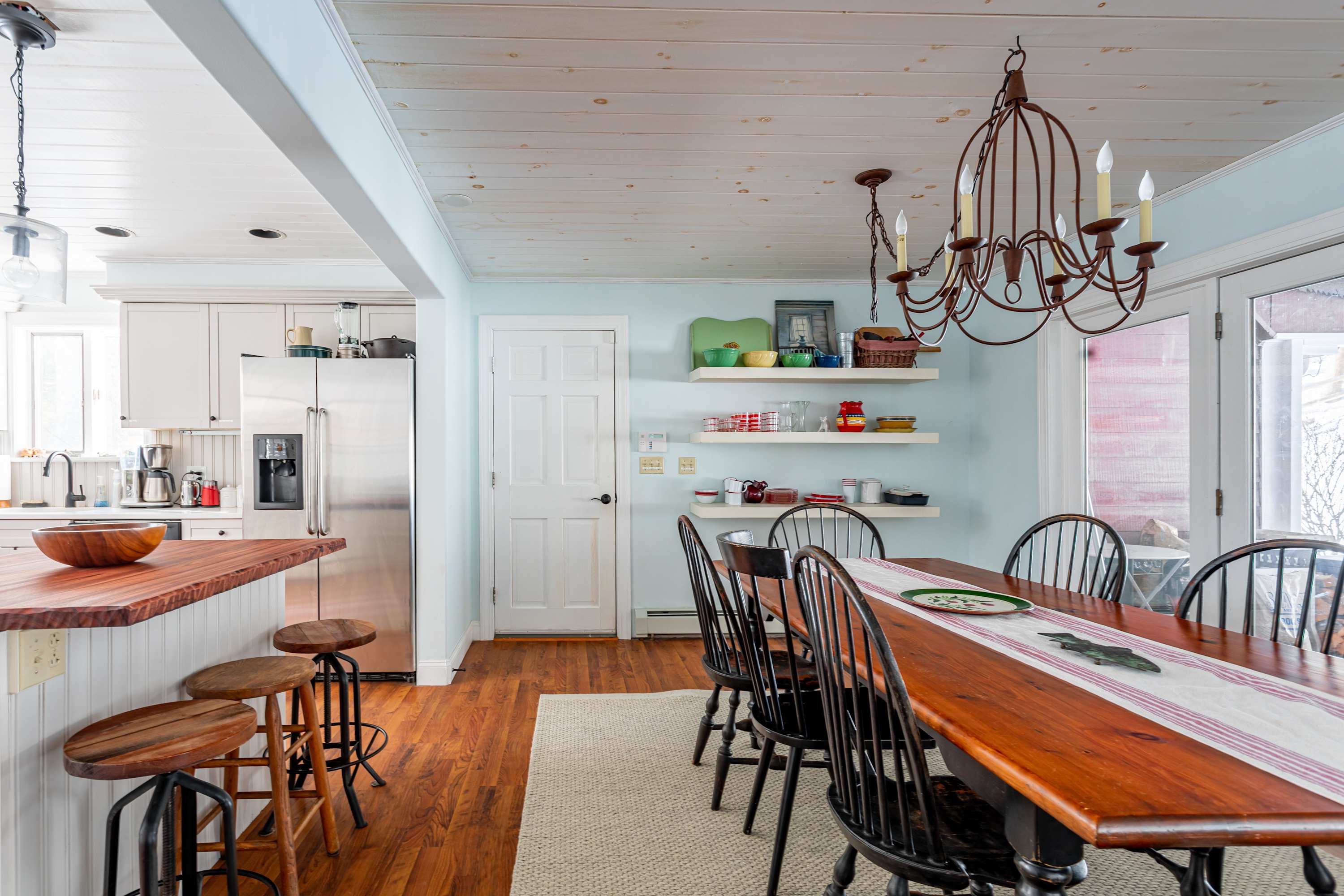 A rustic dining area with a long wooden table, black spindle-back chairs, and a wrought iron chandelier, connected to a white kitchen with a wooden counter and bar stools.