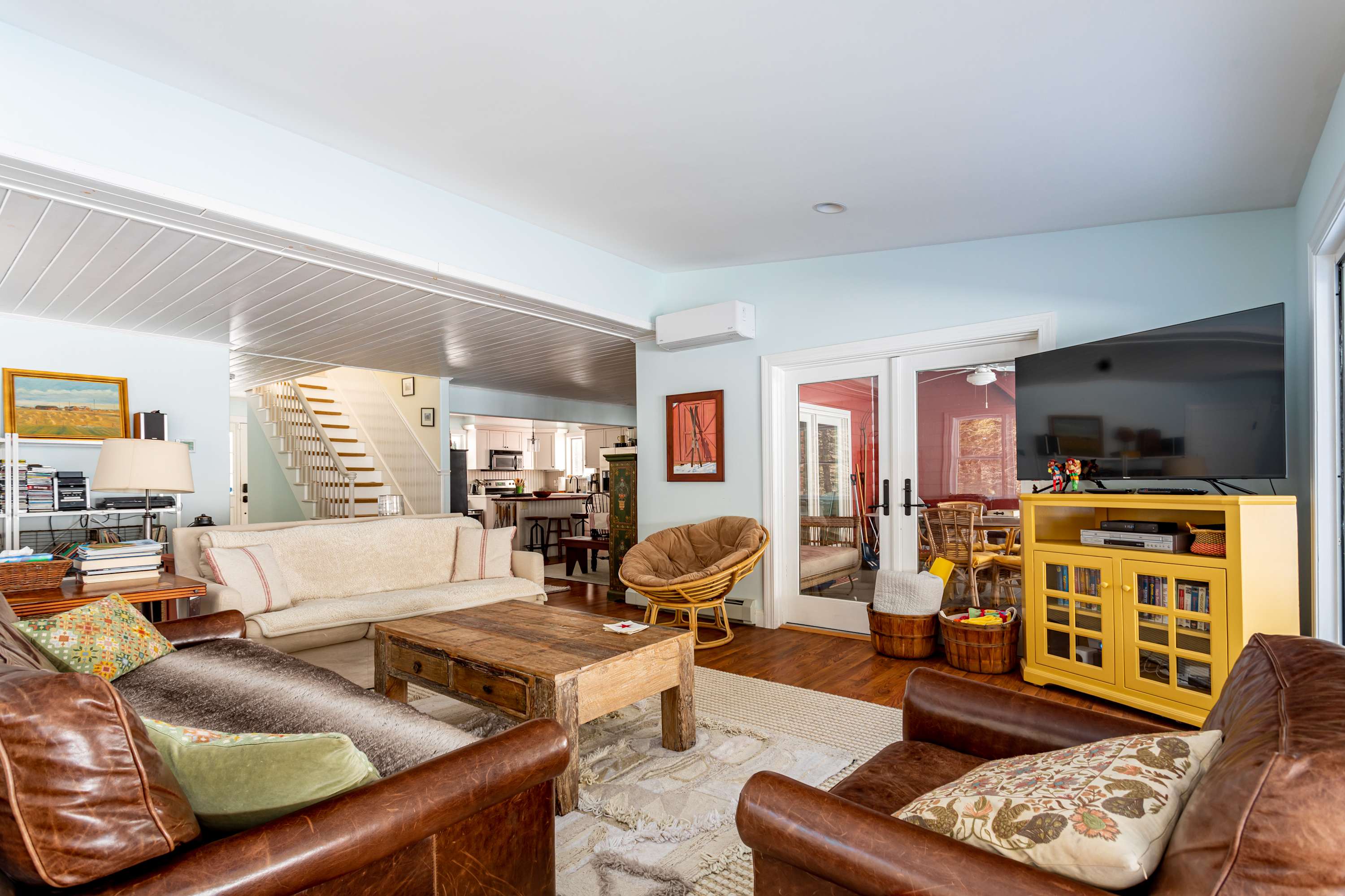 A spacious living room with light blue walls and hardwood floors, featuring a brown leather sofa, a cream fabric sofa, a rustic wooden coffee table, and a large TV, with a view into the dining area and kitchen.