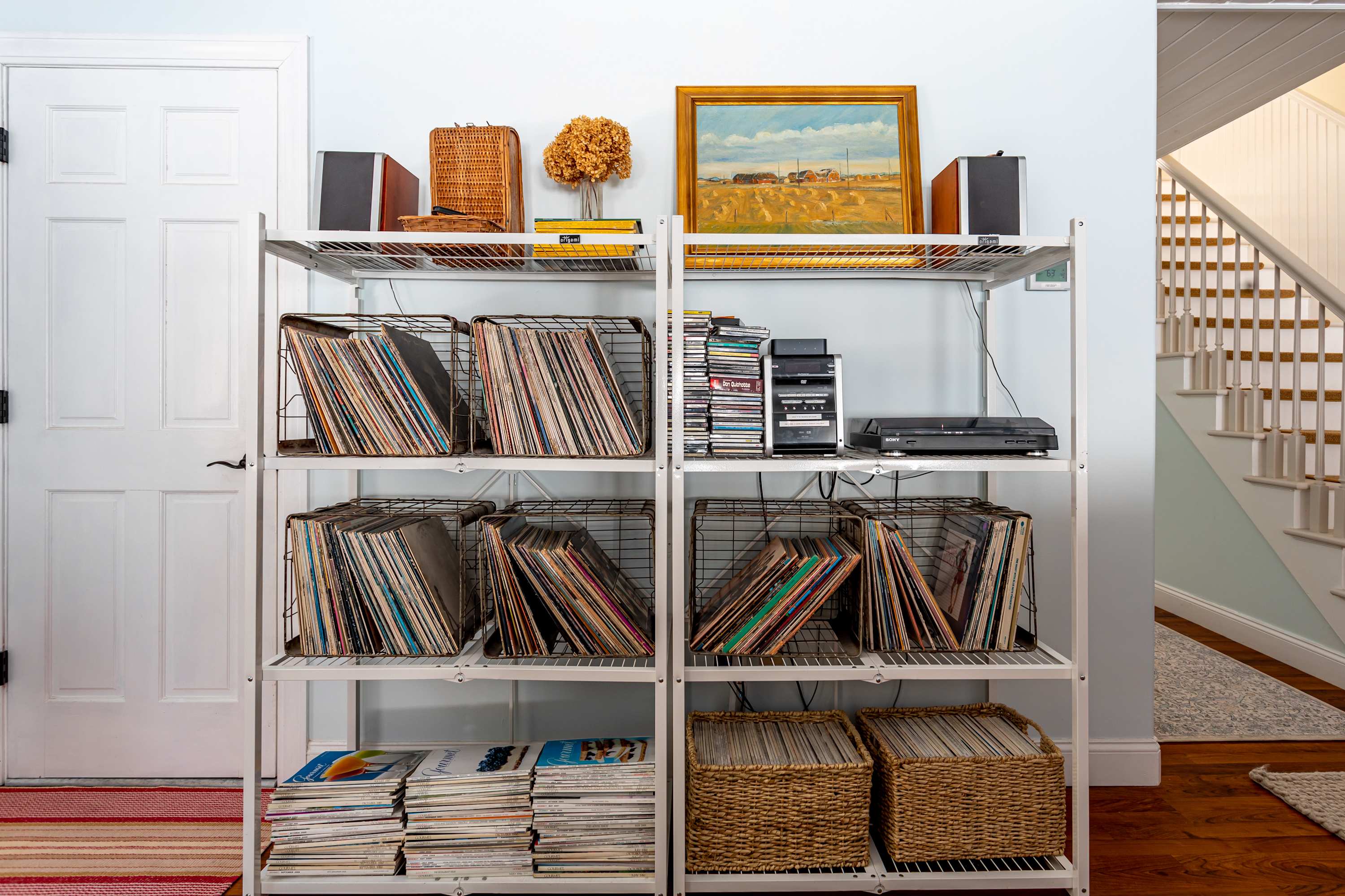 Hallway area featuring a metal shelving unit filled with vinyl records and audio equipment, next to a white door and a wooden staircase with a white railing.