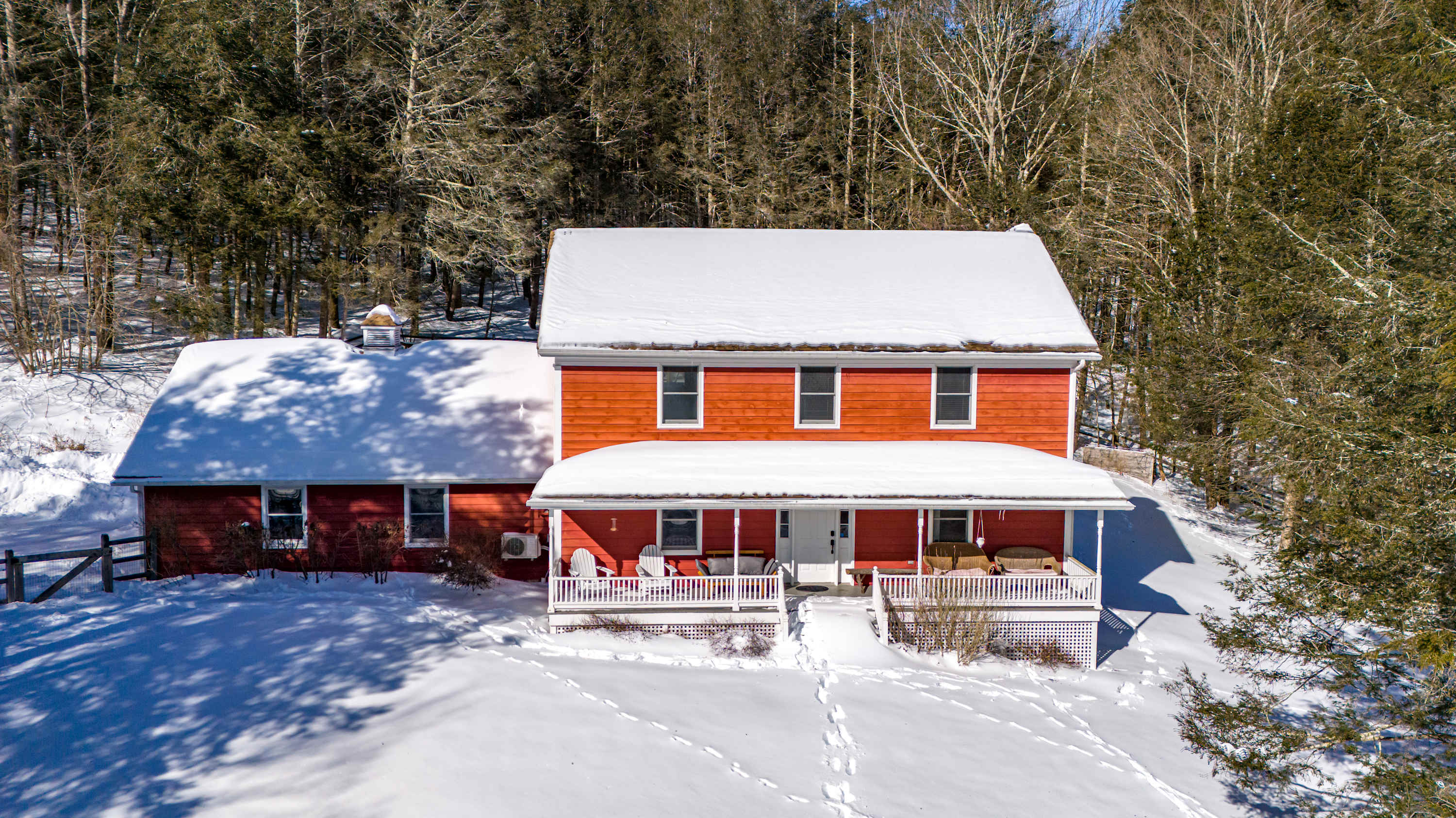 An elevated view captures the red-sided house with its snow-covered roof and wide front porch, surrounded by a pristine blanket of snow and dense winter woods. Footprints trace a path to the entrance, highlighting the serene, secluded setting.