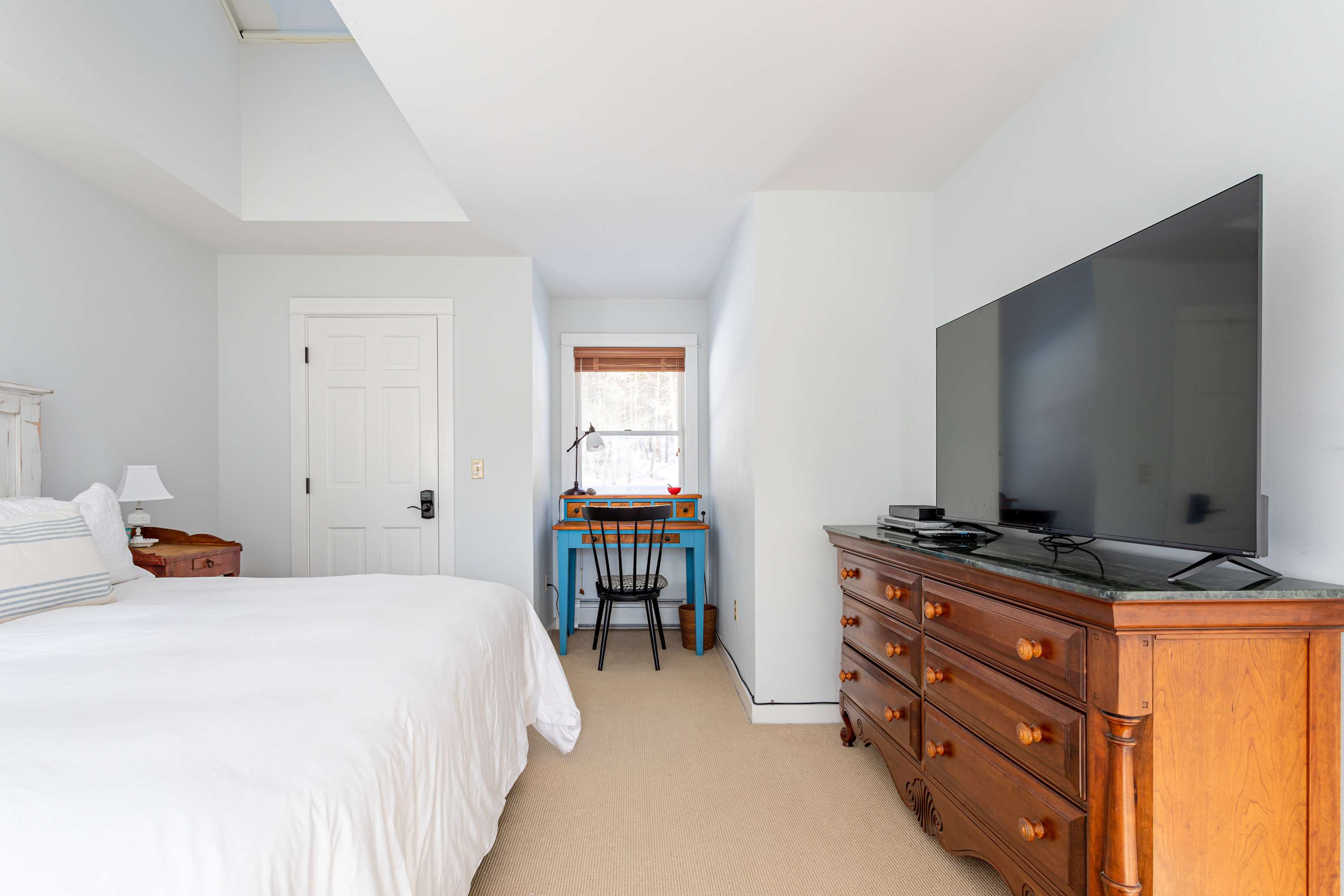 A spacious bedroom featuring a large bed with white bedding, a wooden dresser with a flat-screen TV, and a small blue desk with a chair by a window, all on a light carpeted floor.