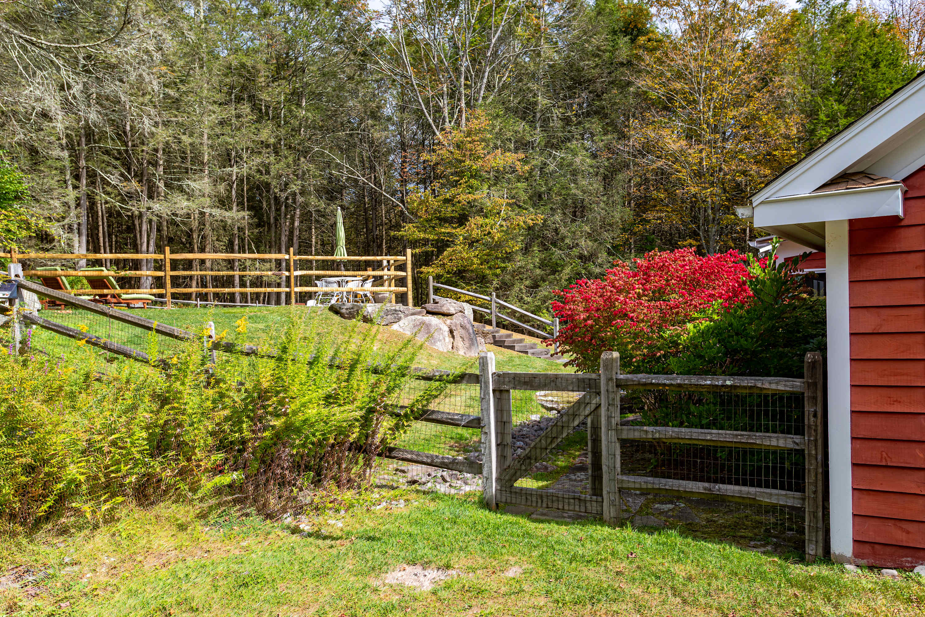 A picturesque view of the property's green yard with a rustic wooden fence and gate, leading up to a stone path and the red-sided house. Vibrant red bushes add a pop of color against the natural backdrop of trees and rocks.