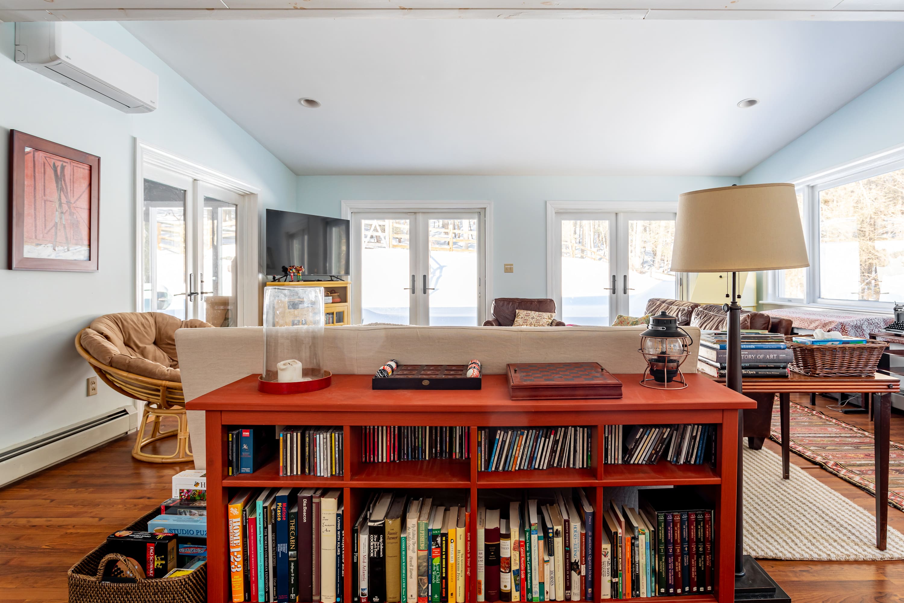 A view from behind a cream fabric sofa in the living room, showing an orange bookshelf filled with books, a floor lamp, and a glimpse of the brown leather sofa and TV area.