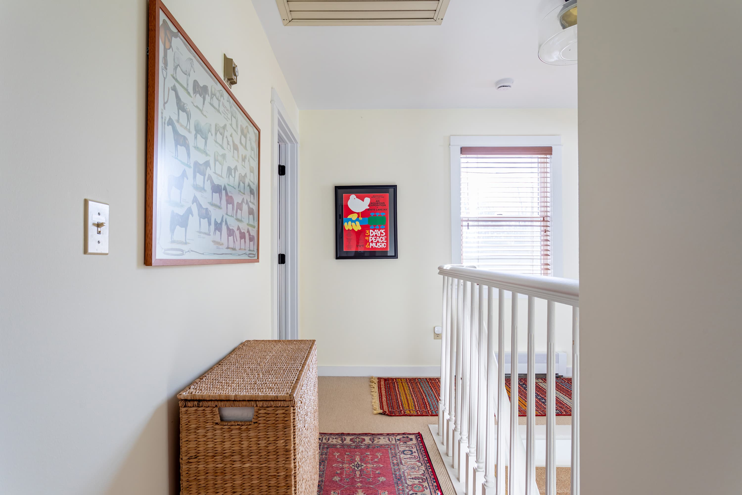 A bright hallway with light yellow walls, a woven storage chest, framed artwork, and a white banister leading to a staircase, with a window providing natural light.