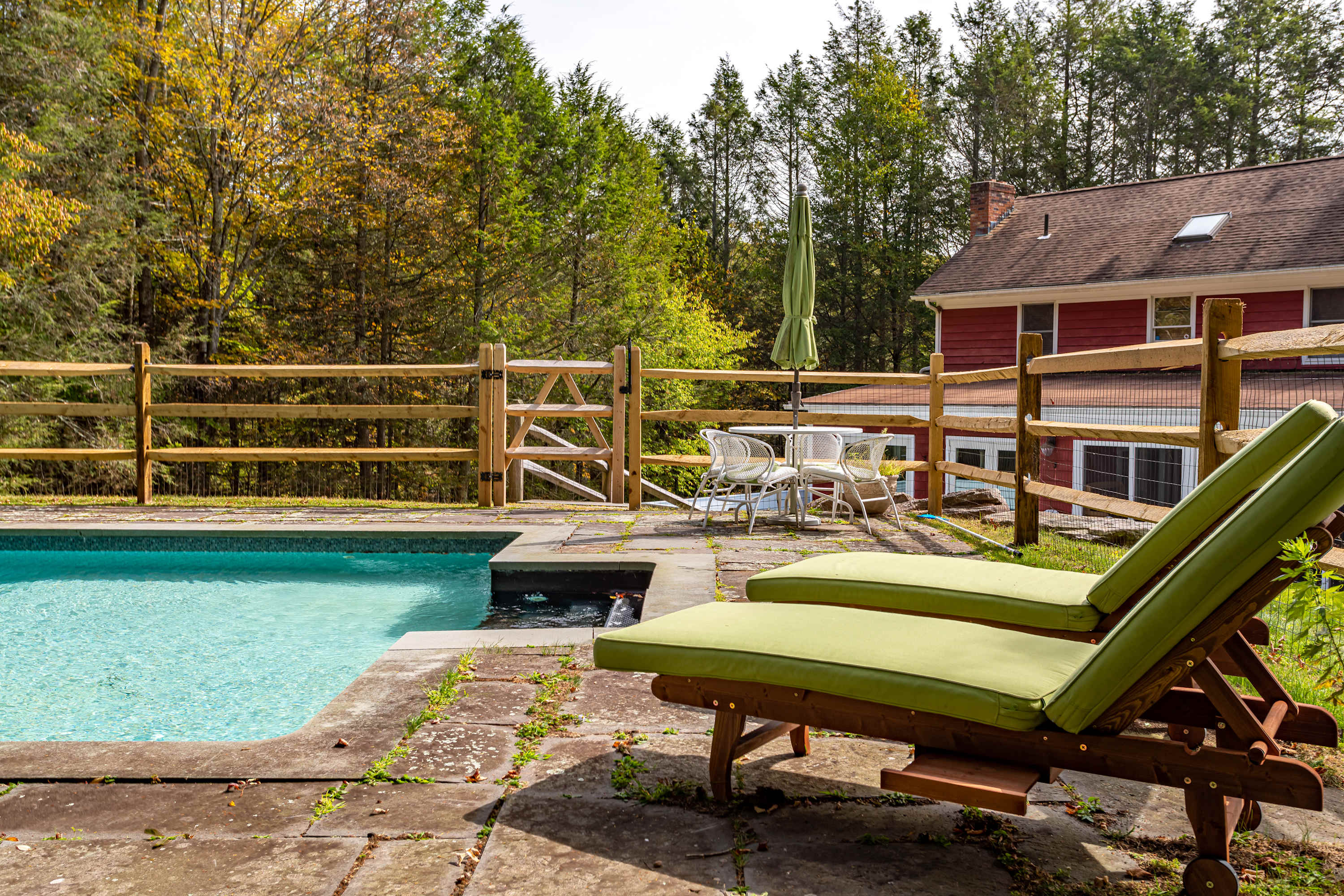 A sparkling blue swimming pool is surrounded by flagstone paving and two green-cushioned lounge chairs, with a wooden split-rail fence enclosing the area. A patio dining set with an umbrella is visible, all set against a backdrop of vibrant autumn trees.
