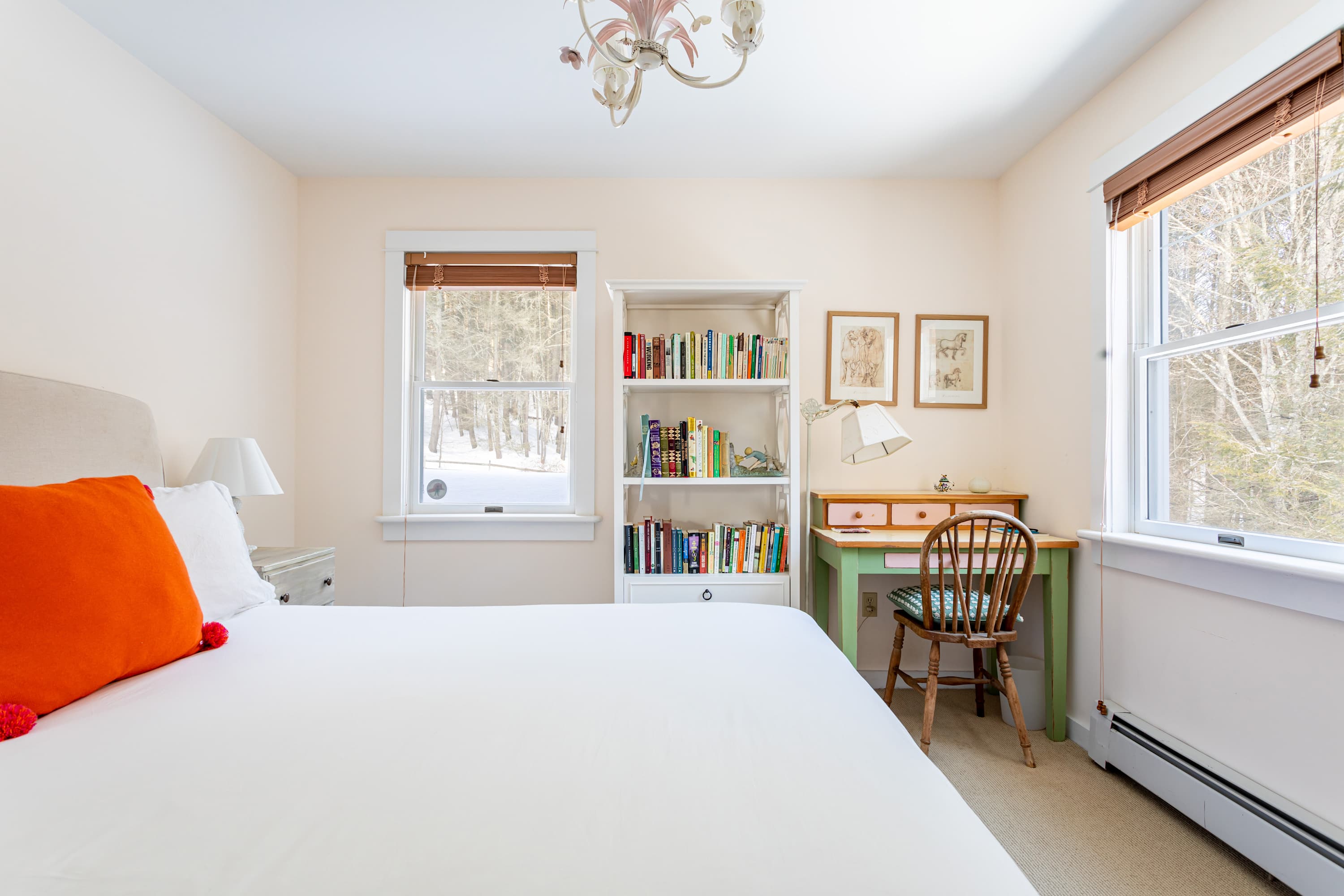 A bright bedroom with a bed featuring white bedding and an orange pillow, a white bookshelf filled with books, and a green desk with a wooden chair, positioned between two windows with wooden blinds.