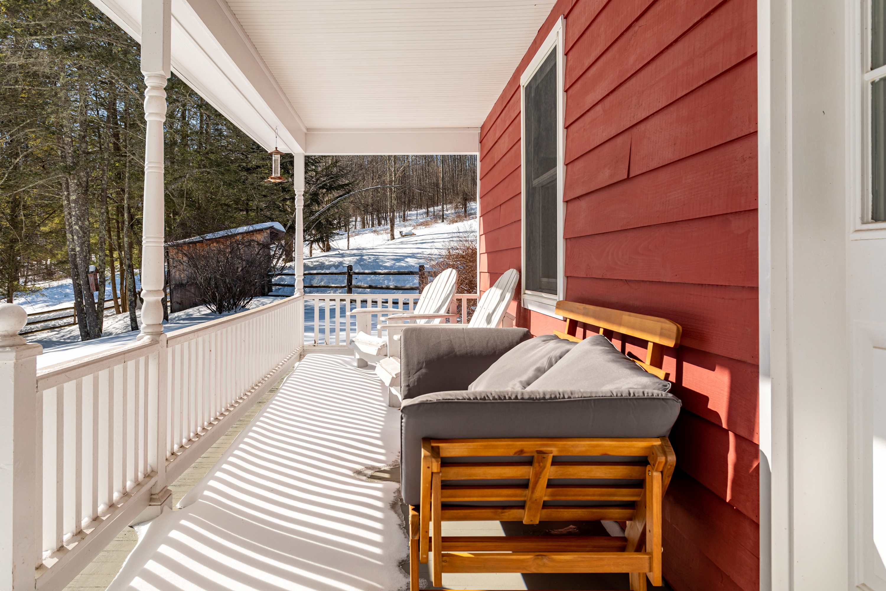 A cozy porch with white railings and a red exterior wall, featuring a gray outdoor sofa and white Adirondack chairs, overlooking a snow-covered wooded landscape.