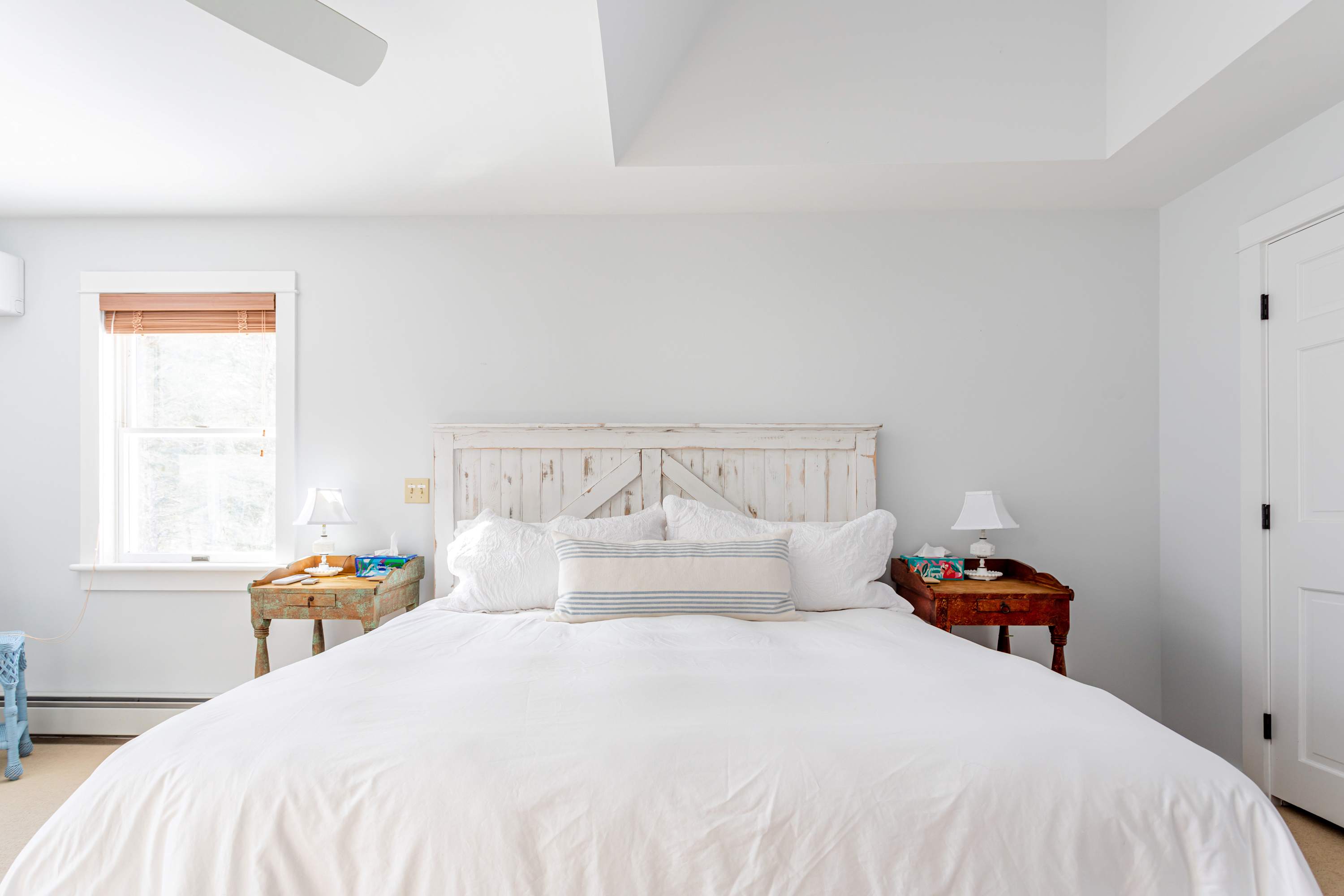 A serene bedroom with light gray walls and a white distressed wooden headboard on a large bed, flanked by two rustic wooden nightstands with lamps, and a window with wooden blinds.
