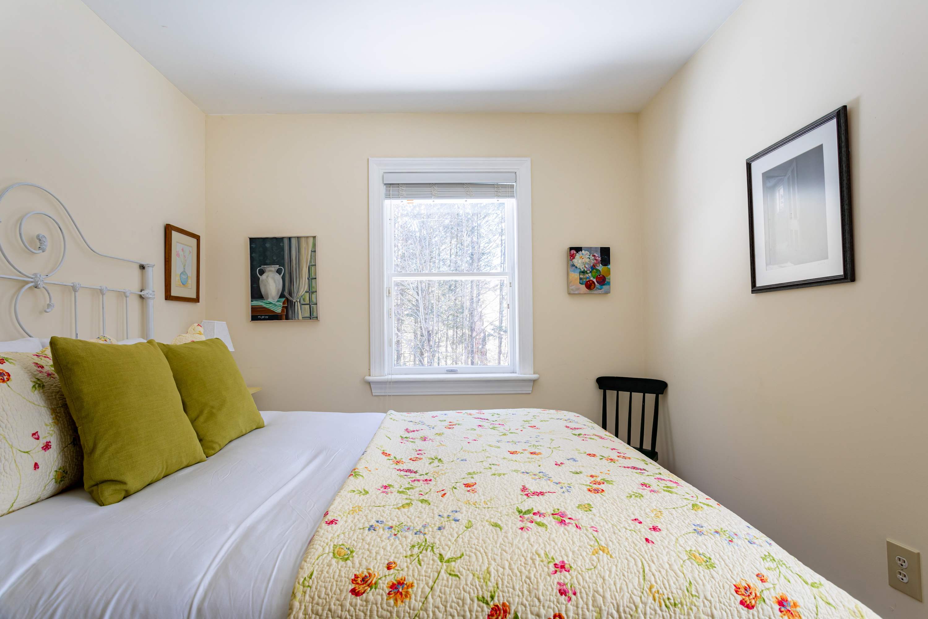 A bright bedroom with light yellow walls, featuring a white metal bed frame dressed with a floral yellow quilt and green throw pillows. A window overlooks a wooded area, flanked by two small framed artworks, with a black wooden chair in the corner.