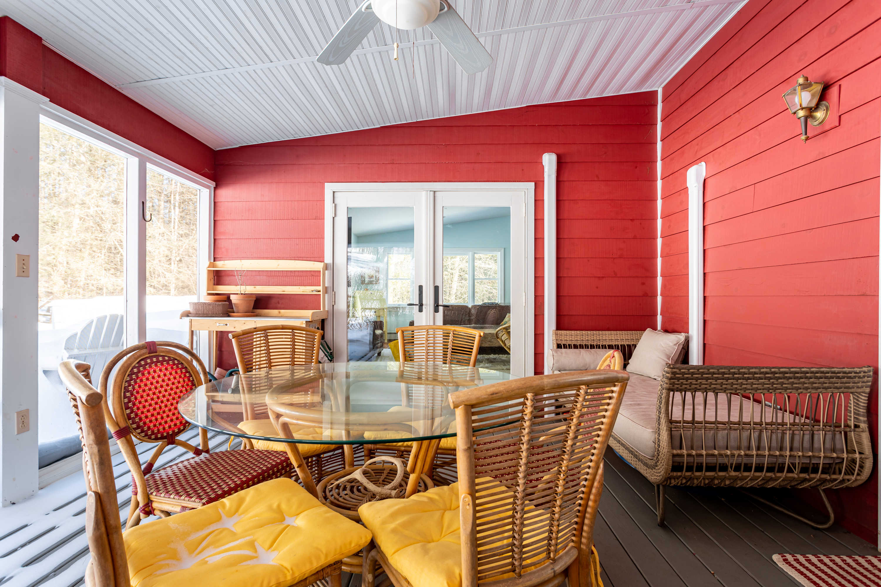Enclosed porch with red shiplap walls, a glass-top table with wicker chairs, and a ceiling fan, offering views of a snowy outdoor landscape through large windows.