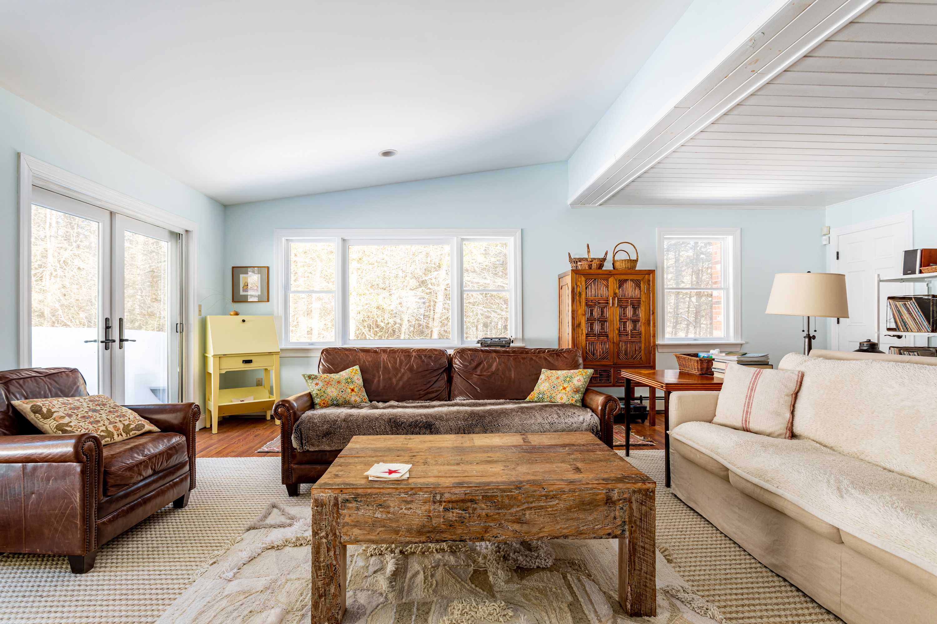 A bright living room with light blue walls and hardwood floors, featuring a brown leather sofa with decorative pillows, a rustic wooden coffee table, and large windows offering a view of the snowy outdoors.