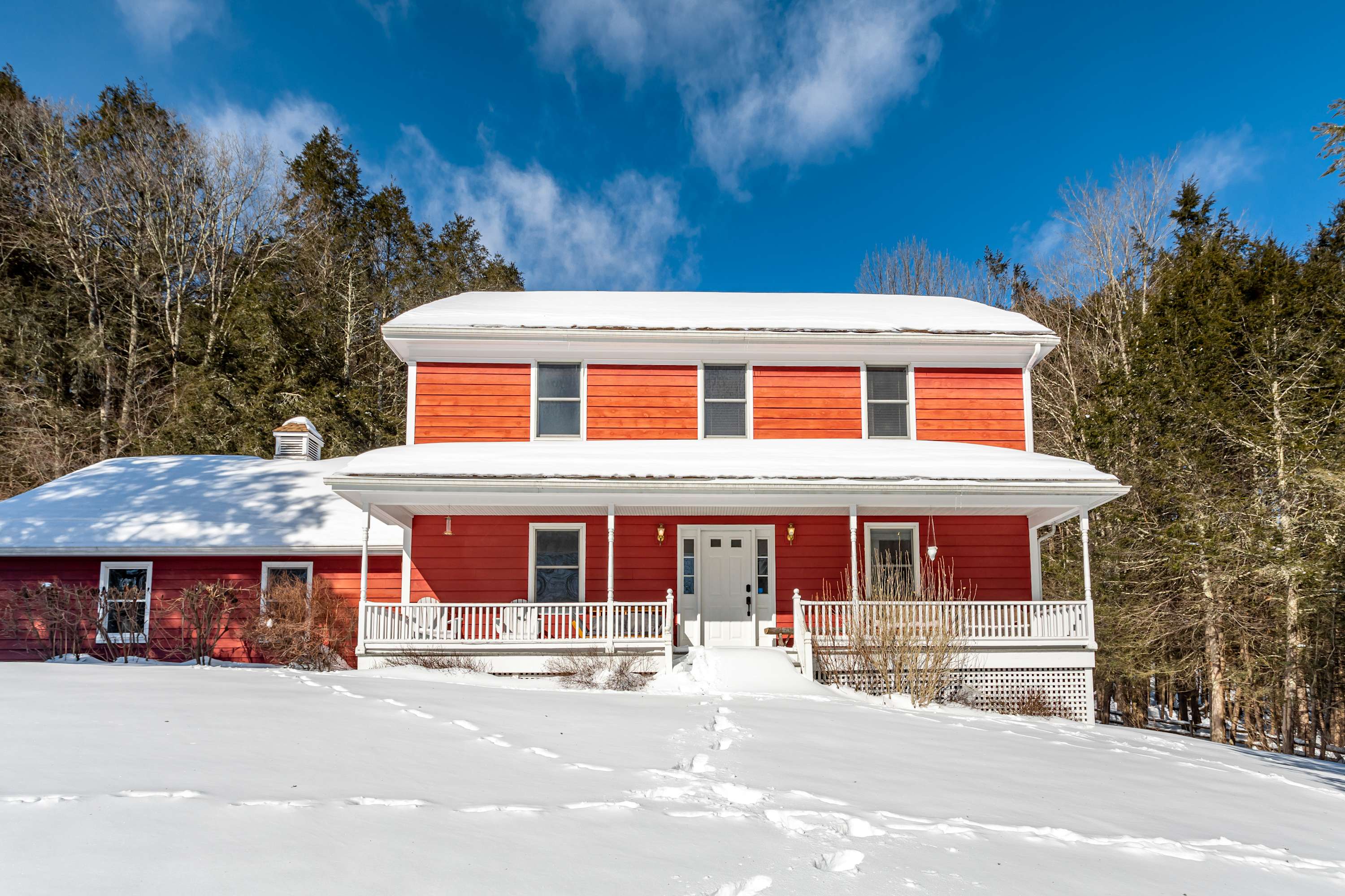 The front facade of the red-sided house is covered in snow, featuring a wide white porch with railings and multiple windows. Footprints lead through the snow to the front door, with a backdrop of bare winter trees and a bright blue sky.