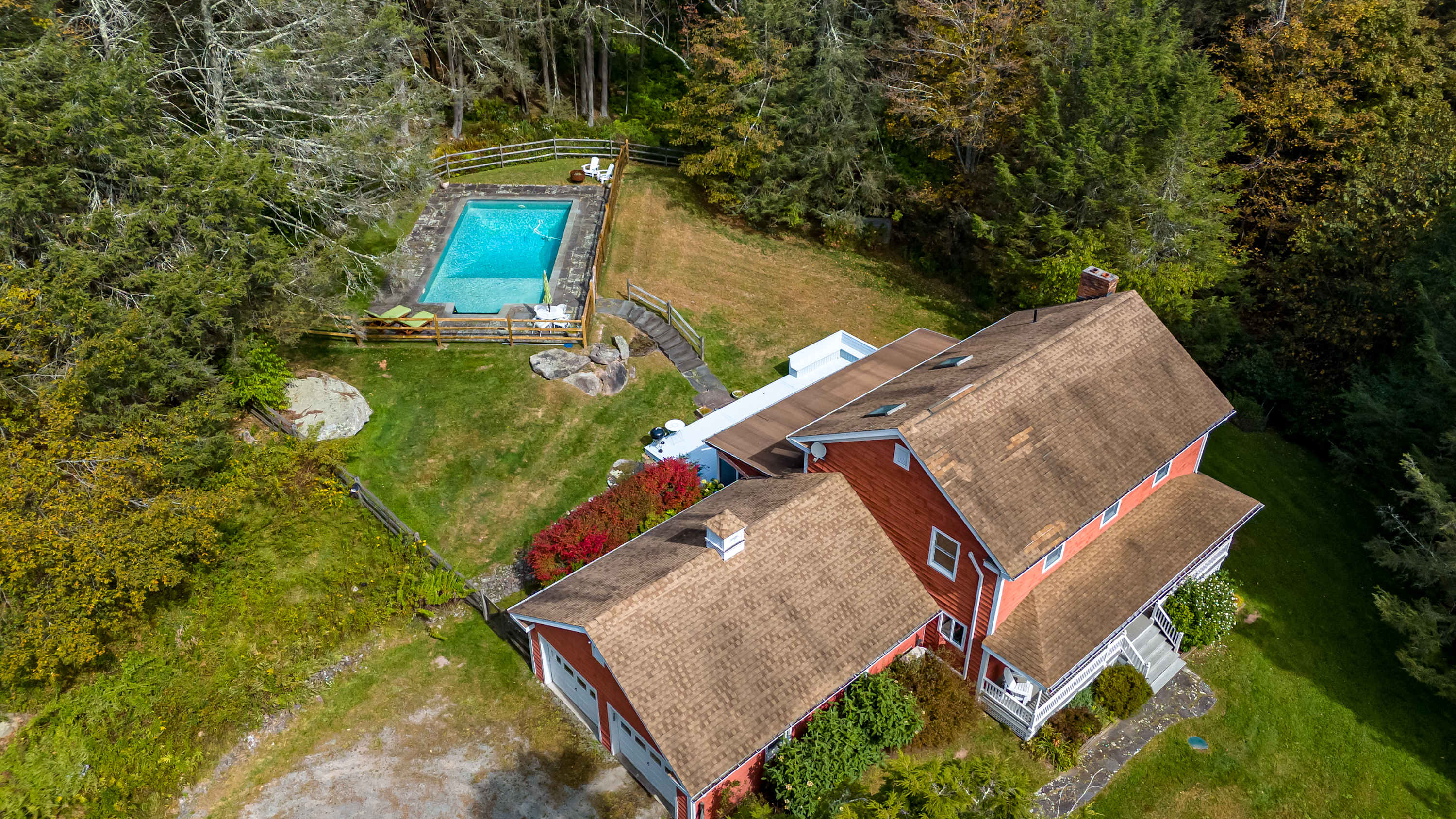 An aerial perspective of the red-sided house with a brown roof, nestled amidst a lush green landscape with dense trees. A rectangular swimming pool with a surrounding patio is visible on the terraced lawn, connected by steps.