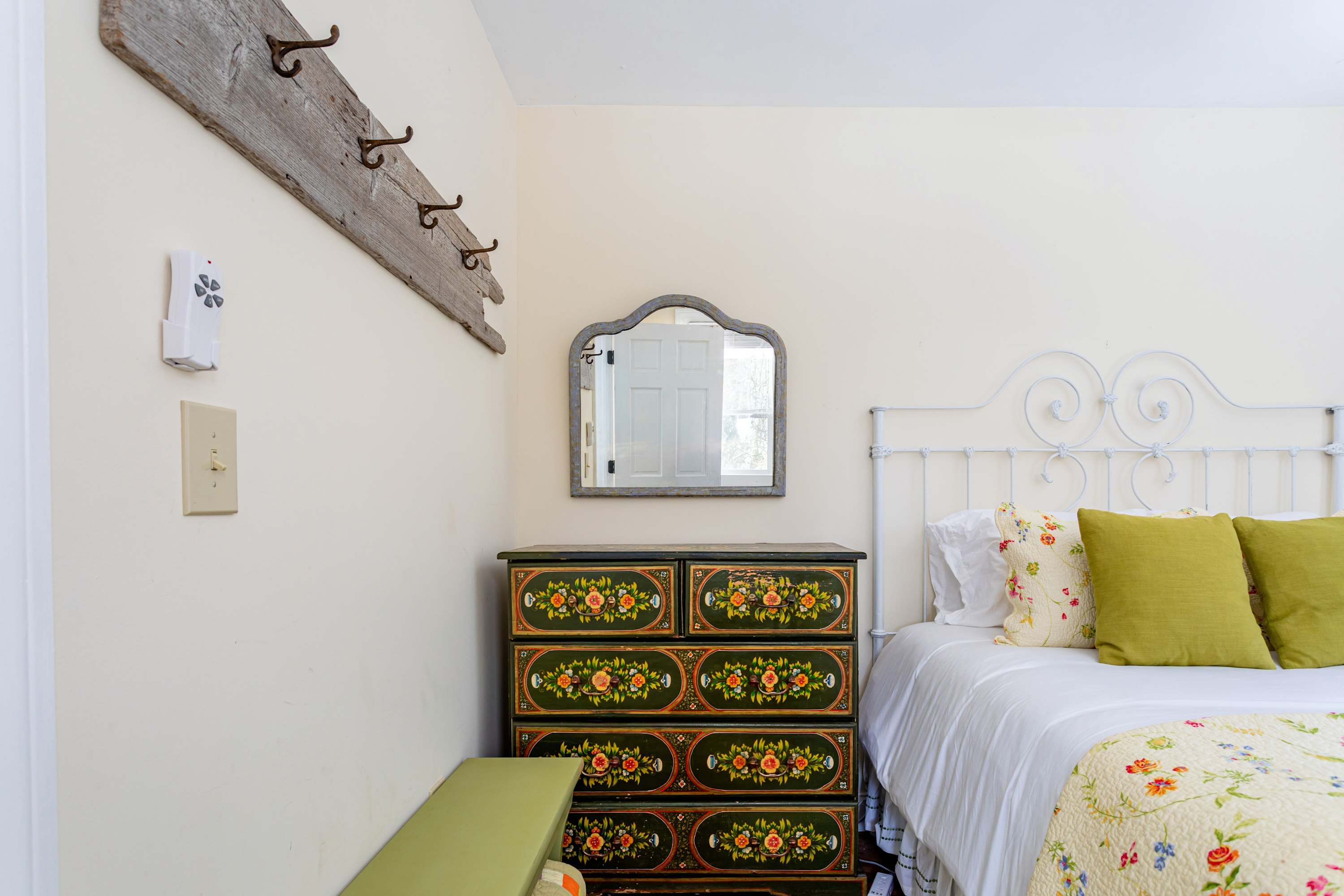 A charming bedroom with a white metal bed frame, a floral quilt, and green pillows, alongside a dark green dresser with intricate floral paintings. A rustic wooden coat rack and a decorative mirror hang on the light-colored wall.