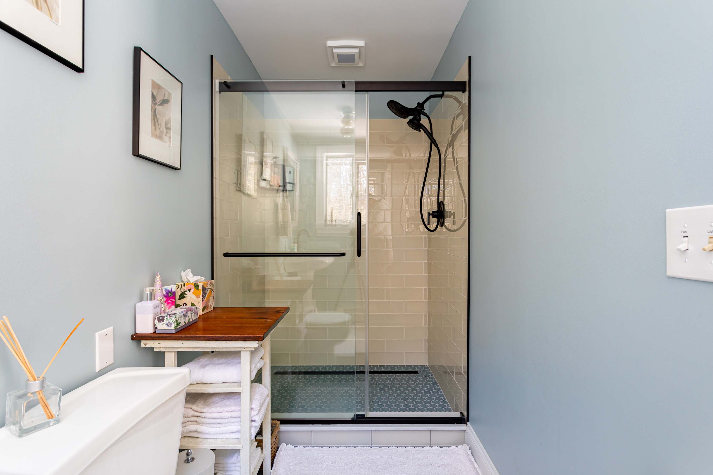 A modern bathroom with light blue walls, featuring a spacious glass-enclosed shower with subway tiles and a black shower head. A white toilet is visible next to a wooden shelving unit holding towels and toiletries.