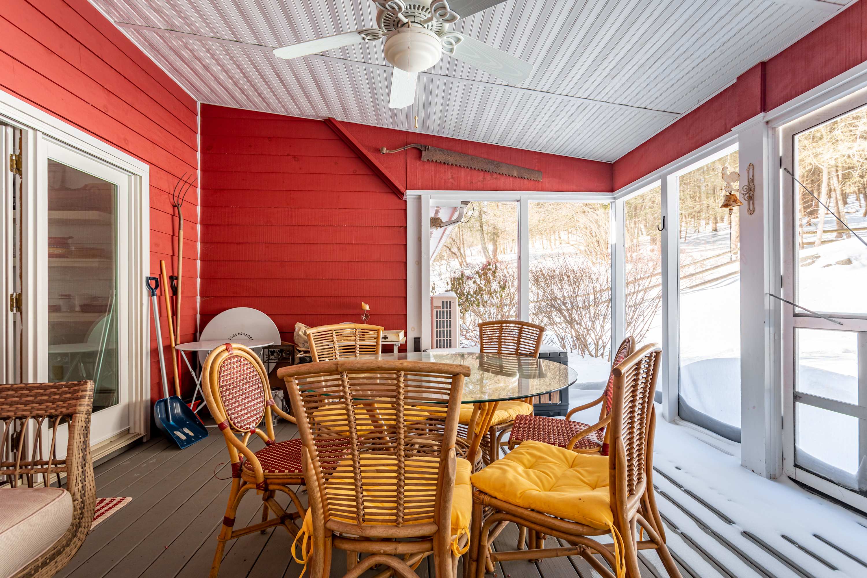 A vibrant screened-in porch with bright red paneled walls and a white ceiling, furnished with a glass-top dining table and six wicker chairs with yellow cushions. Large windows offer views of a snowy outdoor landscape.
