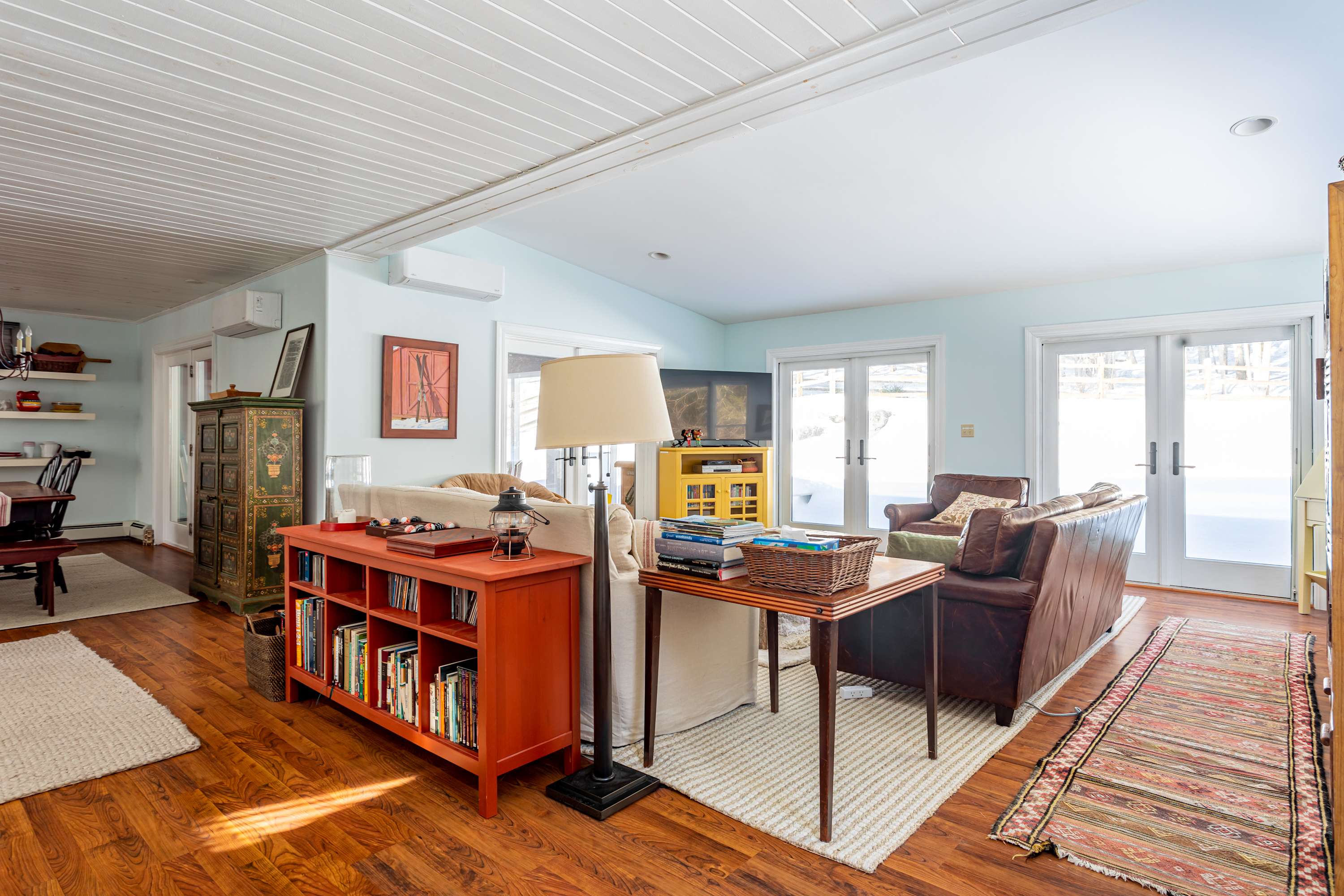 A wide shot of the living room with light blue walls and hardwood floors, showing a brown leather sofa, a cream fabric sofa, a rustic wooden coffee table, and an orange bookshelf, with a view towards the dining area.