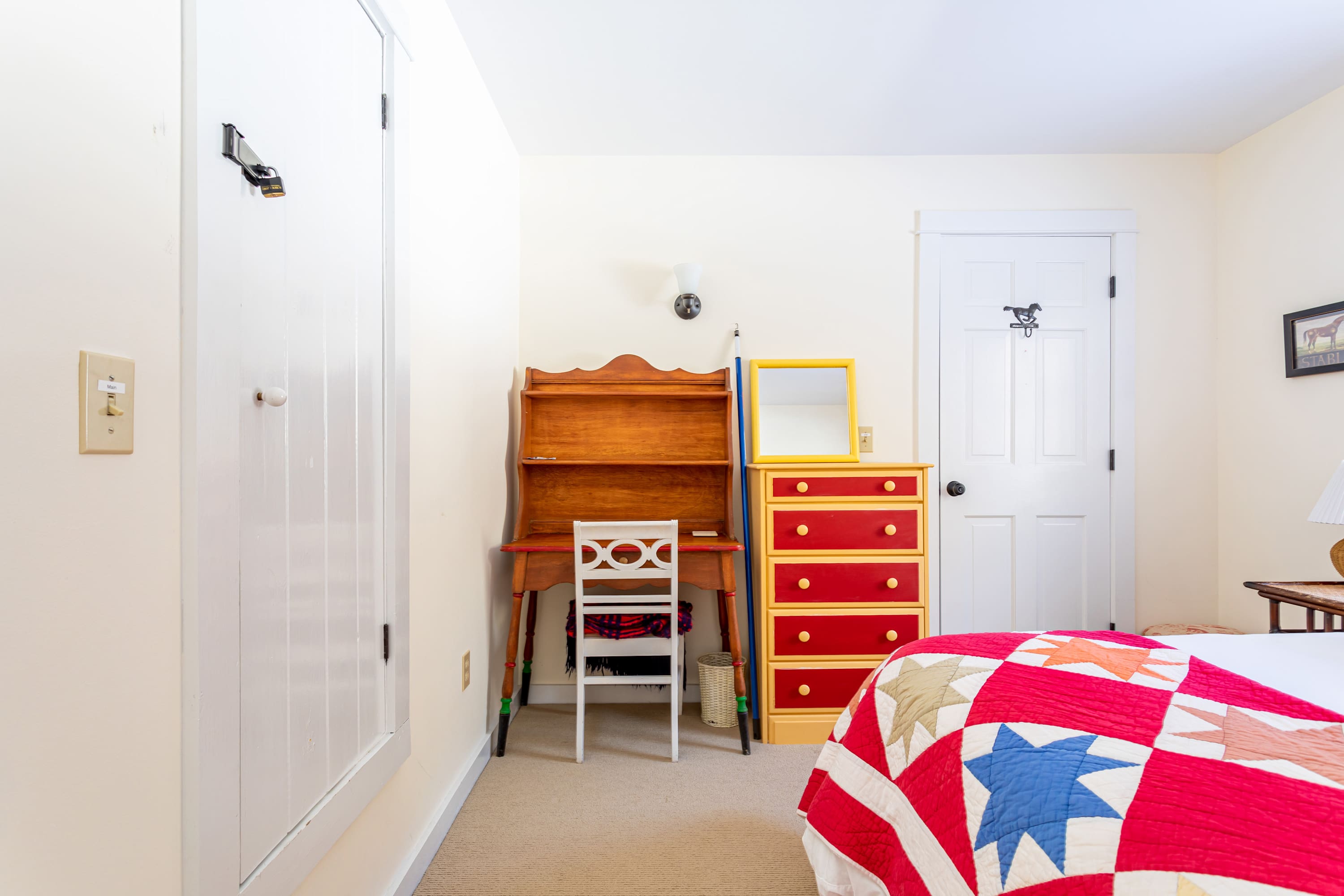 A cozy bedroom featuring a bed with a vibrant red, white, and blue star-patterned quilt, a wooden secretary desk with a white chair, and a bright red dresser with yellow drawers. The room has light yellow walls and a white door with a horse-shaped door knocker.