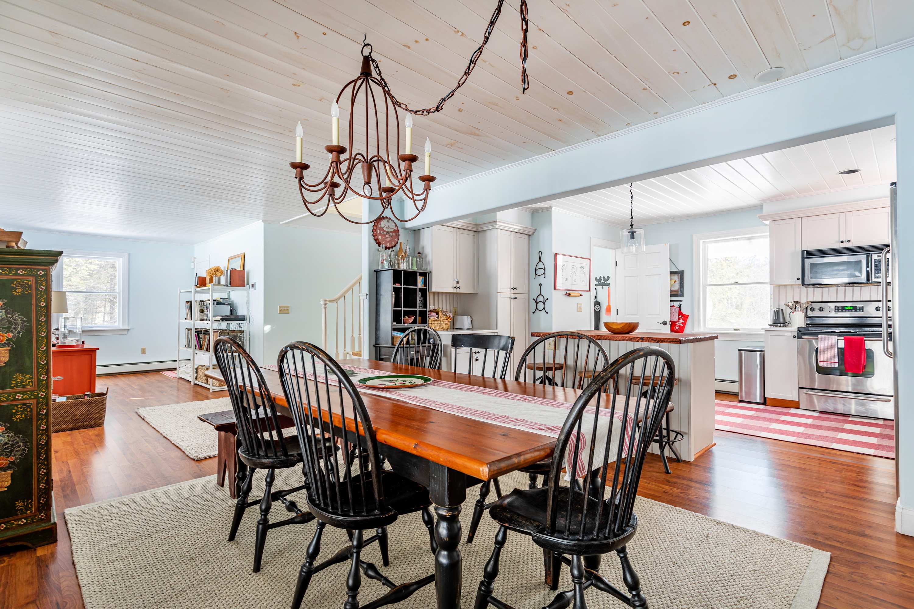 An open-concept dining room featuring a large wooden table with black spindle chairs, a wrought iron chandelier, and a light-colored rug, flowing into a living area and kitchen.