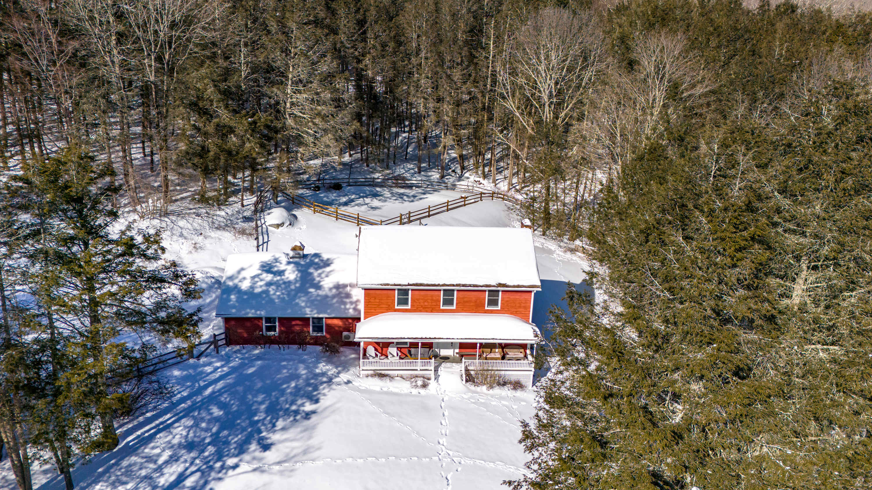 An aerial view of the red-sided house with a snow-covered roof, nestled among bare winter trees and surrounded by a blanket of white snow. Footprints lead up to the front porch, and a wooden fence is visible in the background.