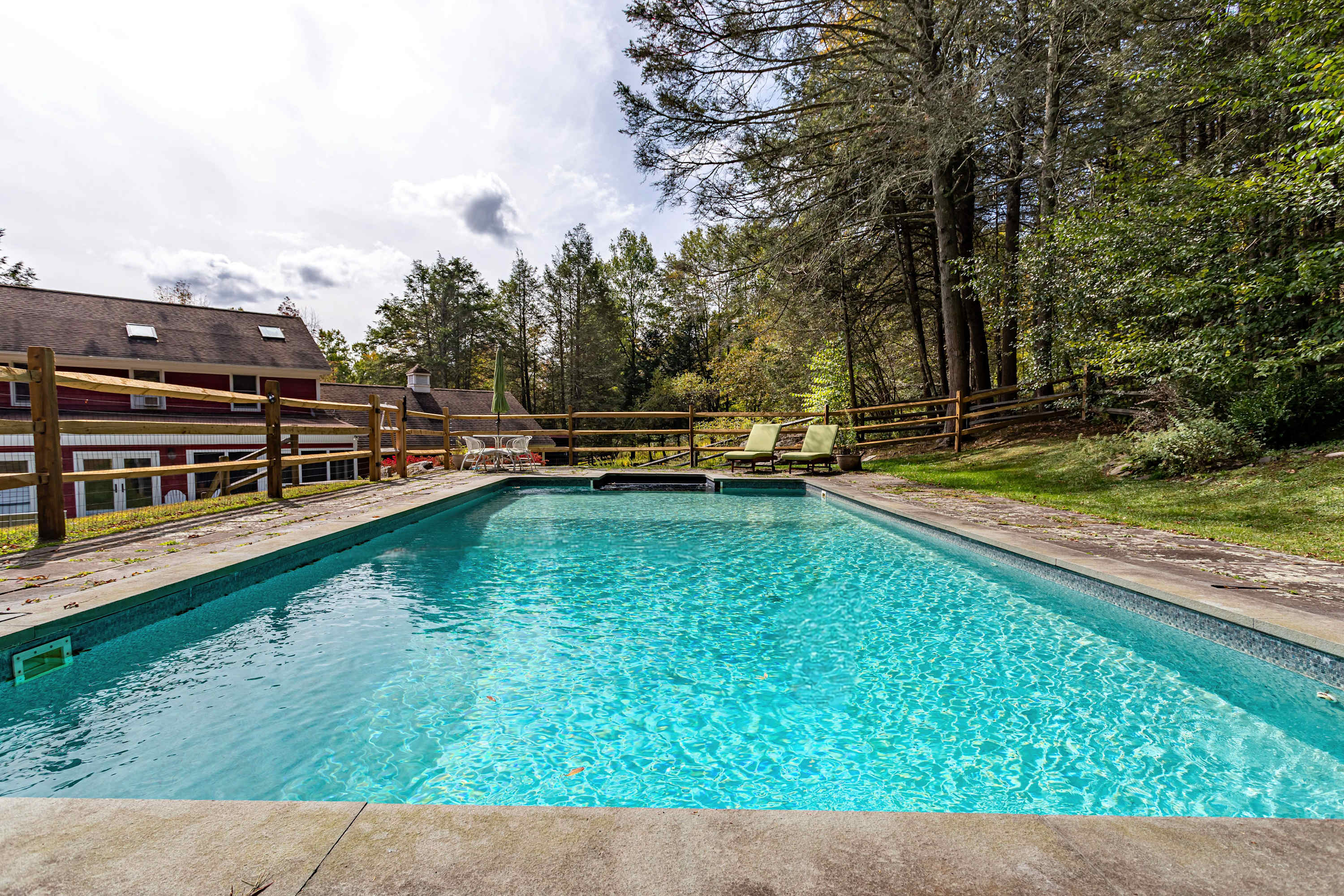 A wide shot of the rectangular swimming pool with vibrant blue water, set within a stone patio and surrounded by a well-maintained green lawn. A wooden fence borders the property, with a dense forest providing a natural backdrop.