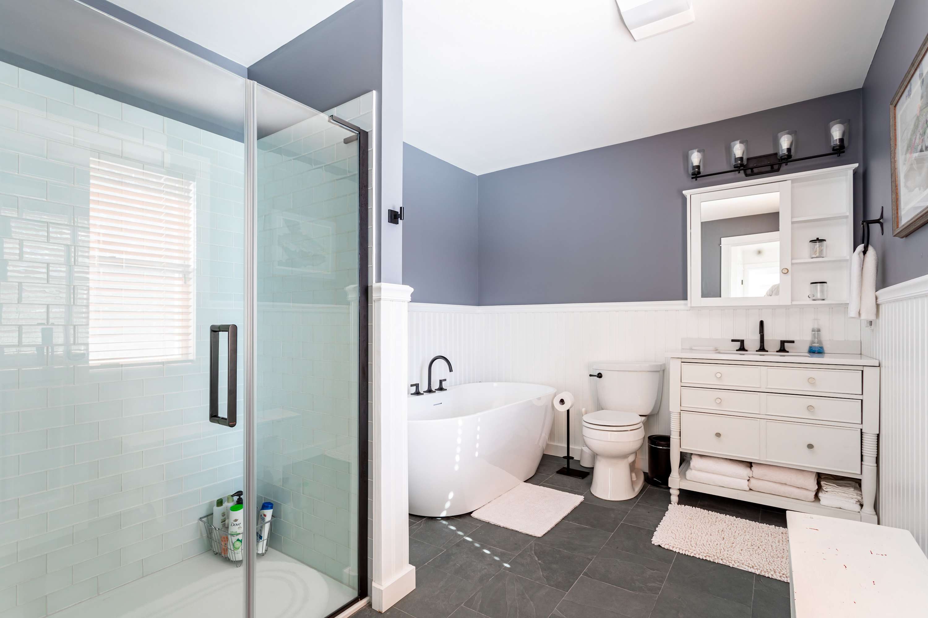 A modern bathroom with a glass-enclosed shower featuring light blue subway tiles, a white freestanding soaking tub, a toilet, and a white vanity with a mirror, all on dark gray floor tiles.