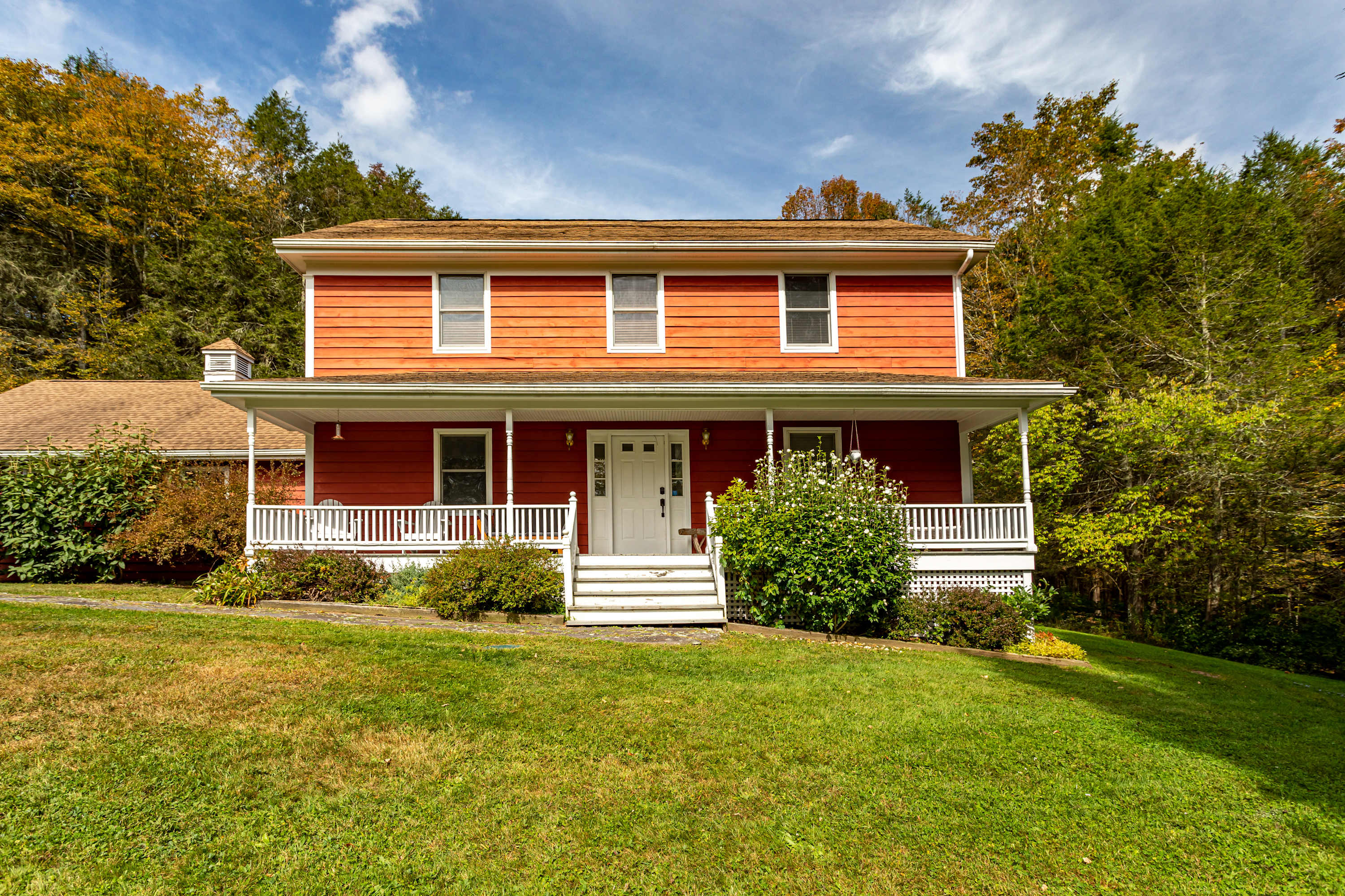 Front view of a two-story red-sided house with a white wrap-around porch, white railings, and steps, surrounded by green grass and mature trees.