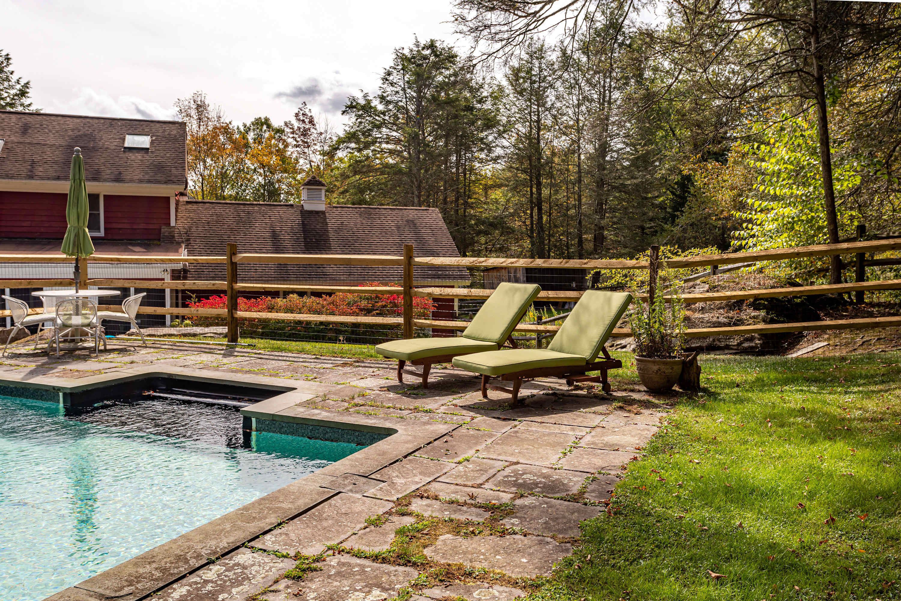 A sunny view of the swimming pool with clear blue water, surrounded by a stone patio and two green lounge chairs. A wooden fence separates the pool area from a grassy slope and distant trees, with the red house visible in the background.