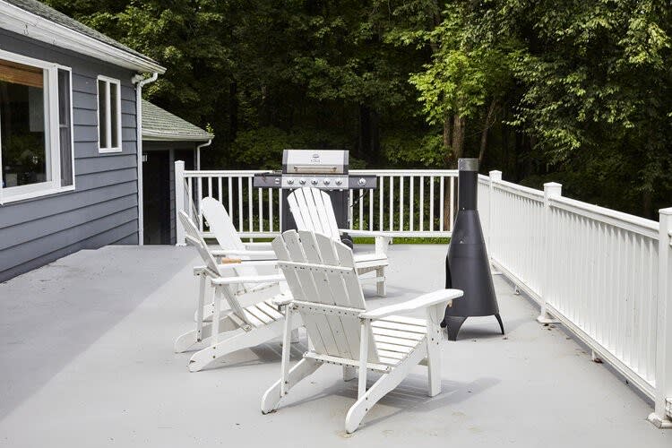 A spacious grey deck features three white Adirondack chairs and a black chiminea, with a BBQ grill positioned against a white railing, overlooking a lush green forest.