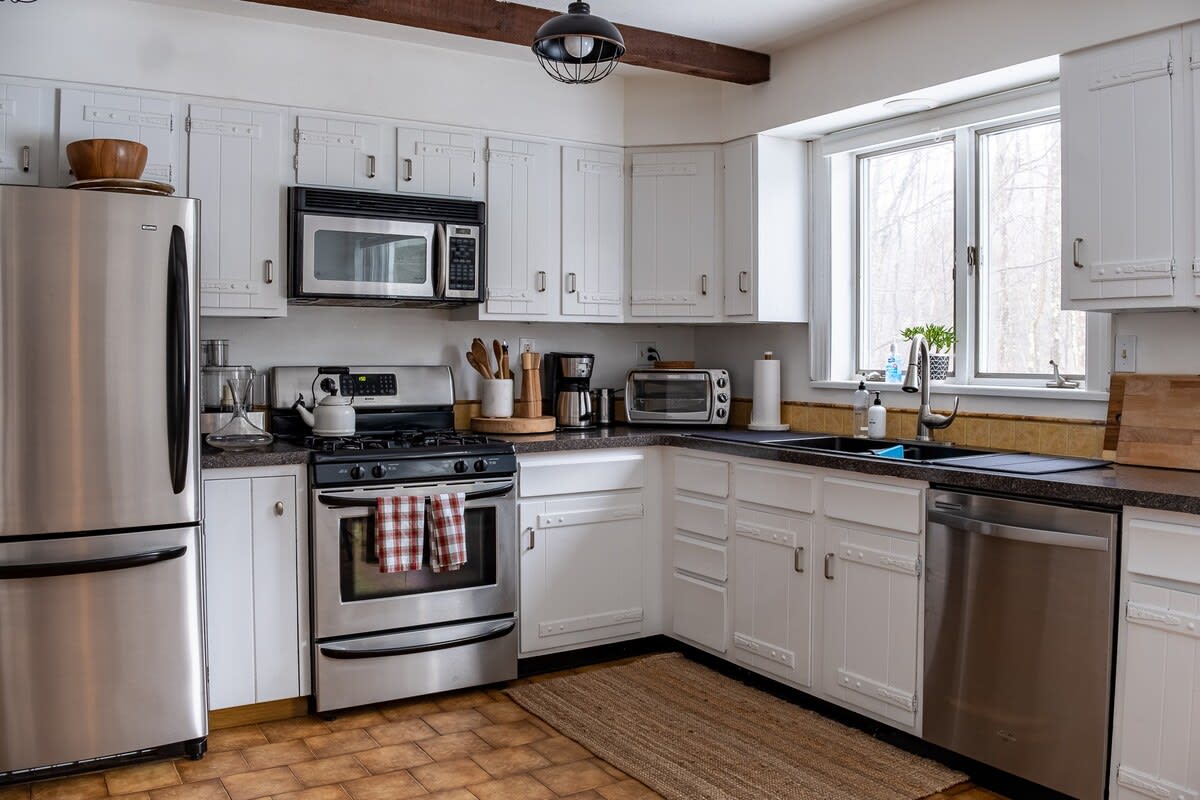 A bright kitchen featuring white shaker-style cabinets, stainless steel appliances including a refrigerator, range, microwave, and dishwasher, dark countertops, and warm-toned tile flooring. A large window overlooks a snowy outdoor scene.