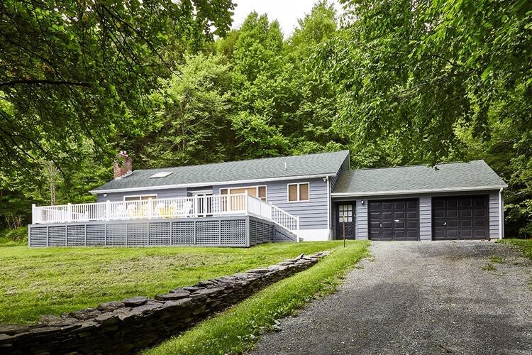 Modern cabin exterior with grey siding and a large grey deck featuring white railings. The house is nestled among lush green trees, with a gravel driveway leading to two dark garage doors.