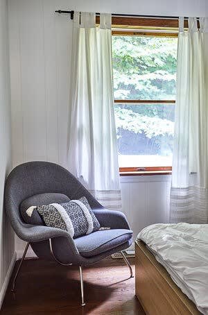 A cozy corner in a bedroom featuring a modern gray upholstered armchair with patterned pillows, positioned next to a window with white curtains. Part of a bed with white bedding is visible to the right.