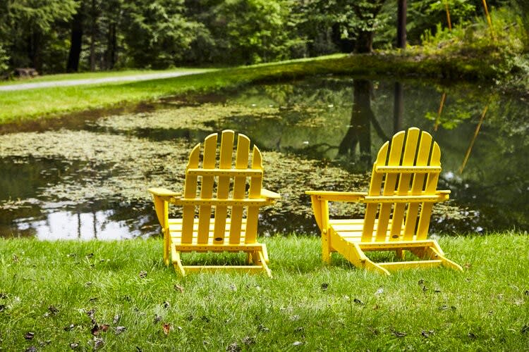 Two bright yellow Adirondack chairs sit on a lush green lawn beside a tranquil pond, reflecting the surrounding trees. The scene evokes a peaceful, natural outdoor setting.